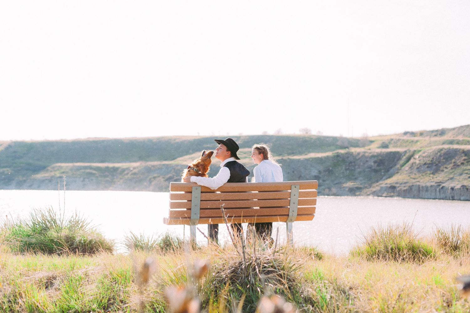 A couple sits on a wooden bench overlooking a scenic coastal landscape with hills and water in the background.