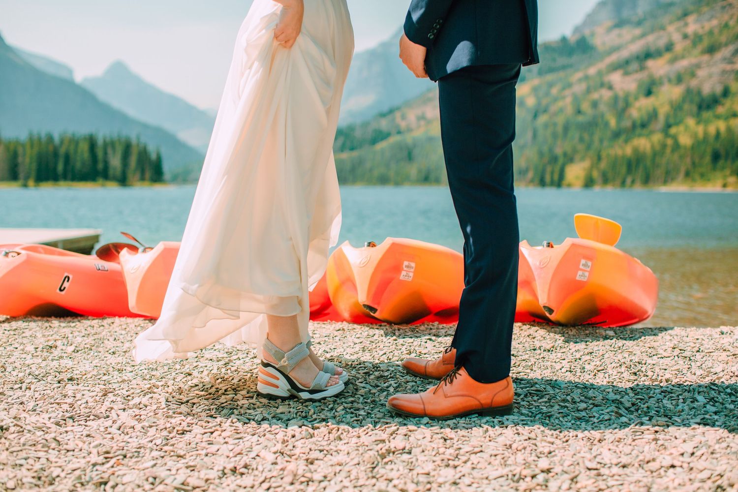 A couple stands at the edge of a lake with mountains and fall foliage in the background during a wedding photoshoot.
