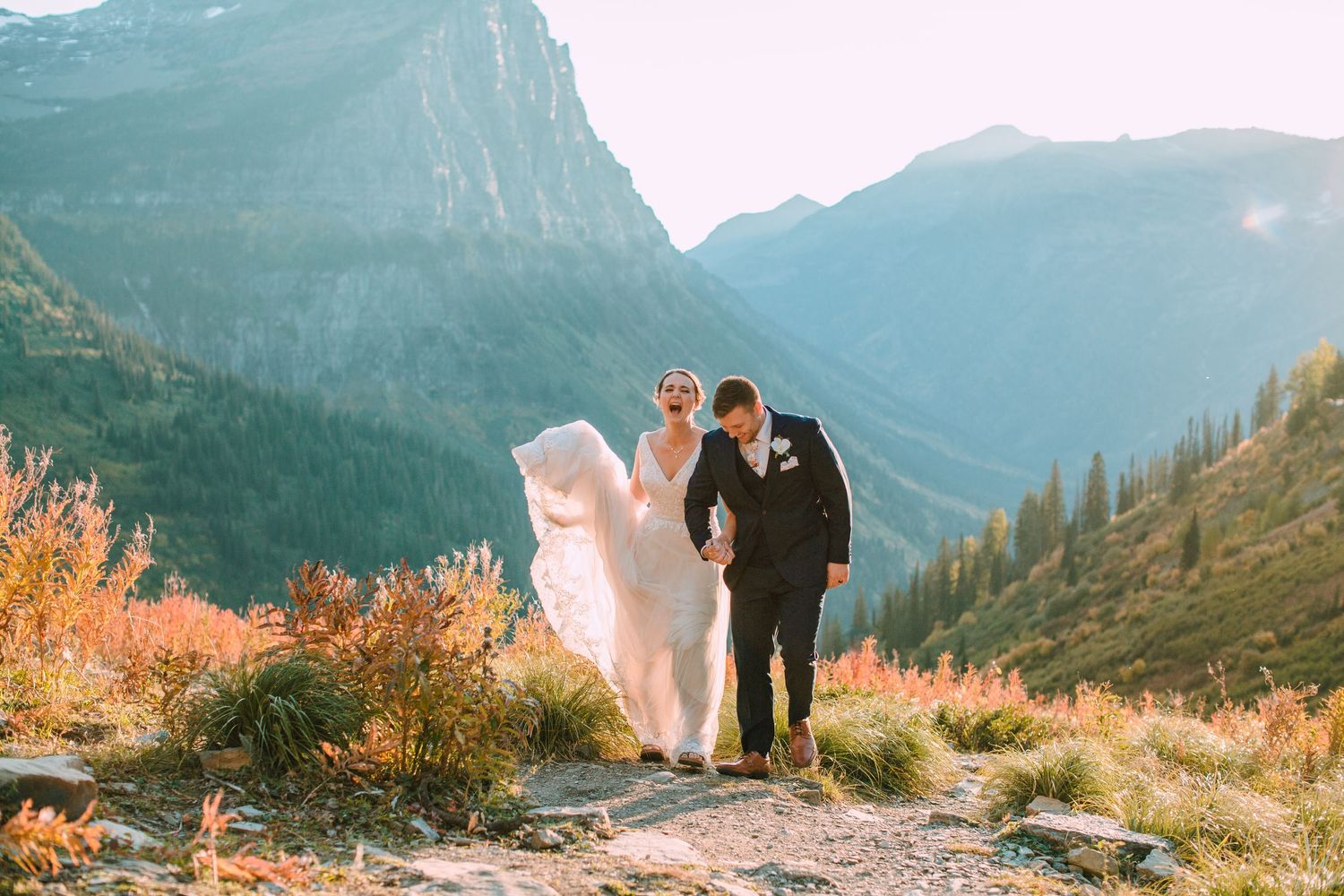 Couple in wedding attire stands on mountain summit with dramatic valley view and autumn foliage in Glacier National Park.