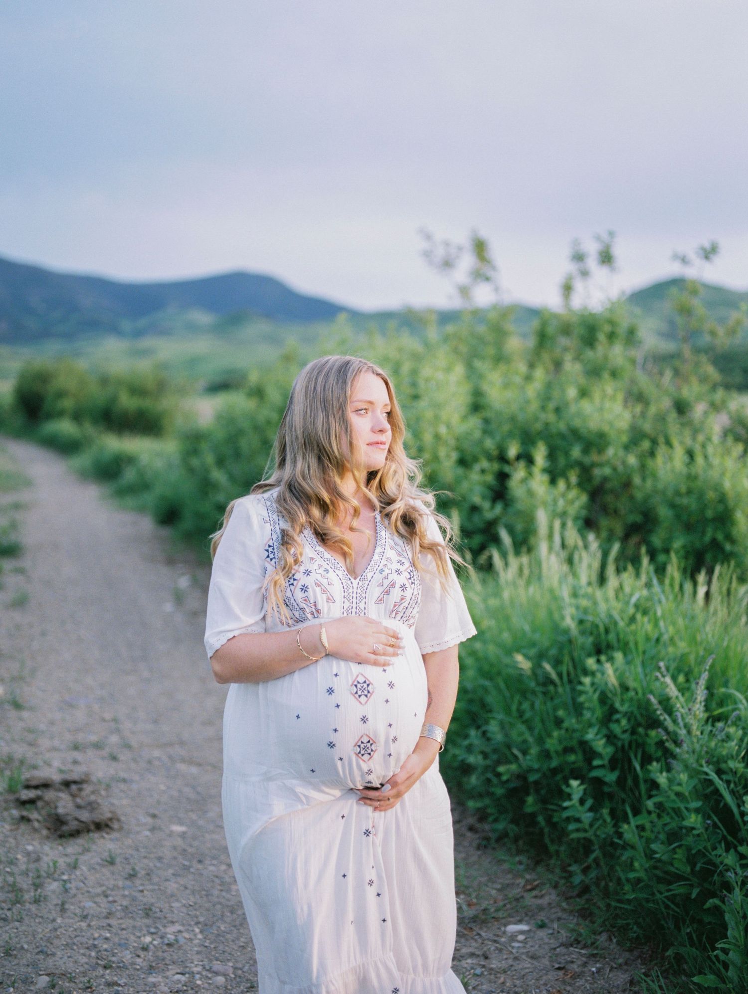 A woman in a flowing white dress stands on a nature trail surrounded by green mountains and wildflowers.