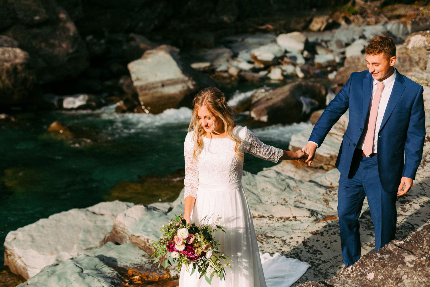 A bride in white dress and groom in blue suit walk along rocky coastal cliffs.