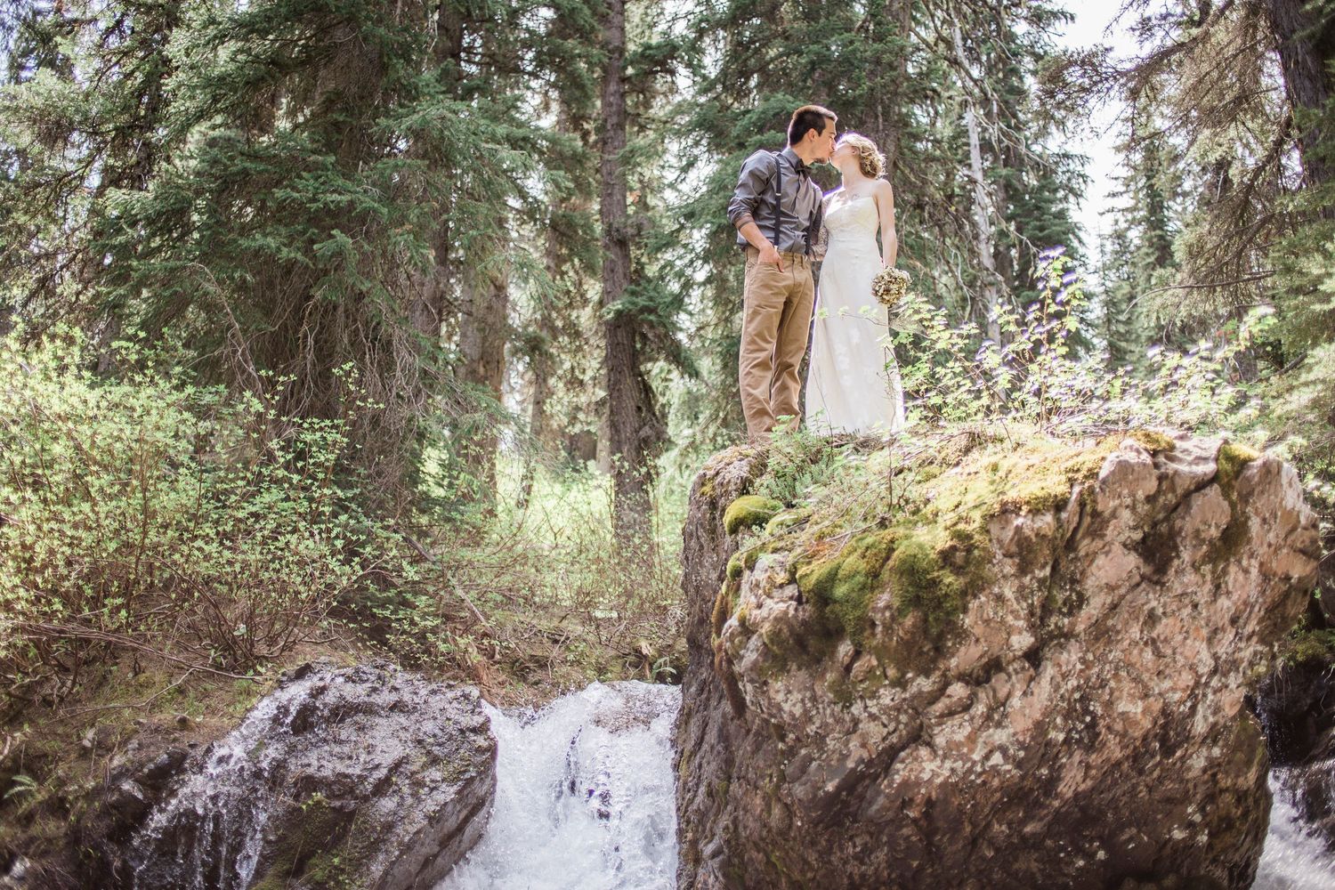 A couple poses in front of a cascading waterfall surrounded by lush green forest during a summer photo shoot.