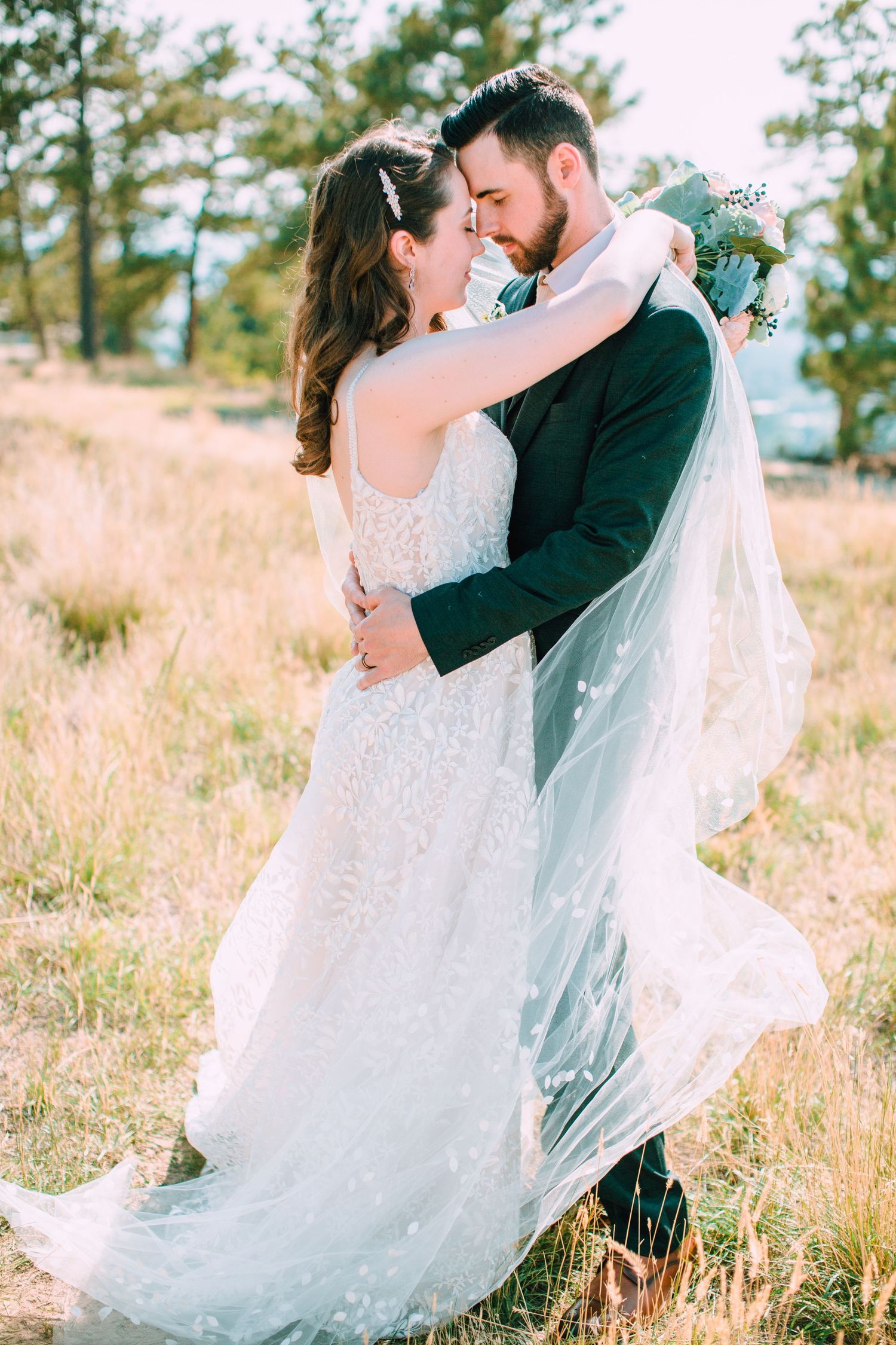 Romantic wedding couple embraces in golden field as bride's white veil flows in gentle breeze.