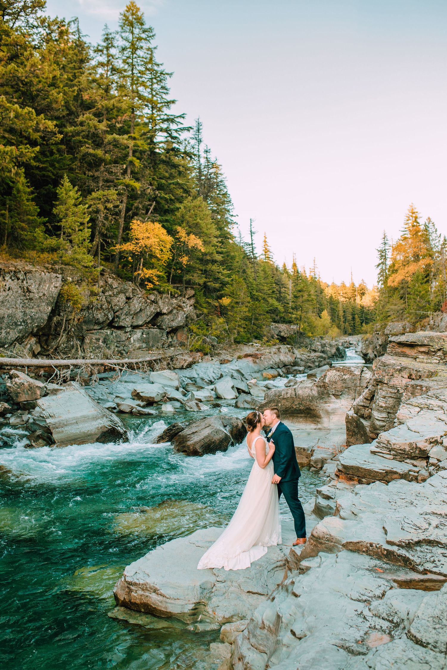 Couple in wedding attire standing on rocky cliffs beside turquoise rushing river in mountain forest landscape at sunset.