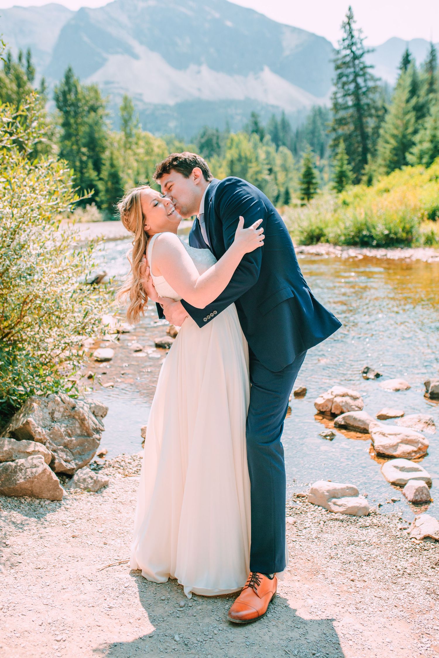 A romantic couple shares a tender moment by a mountain stream surrounded by evergreen trees and rocky terrain.