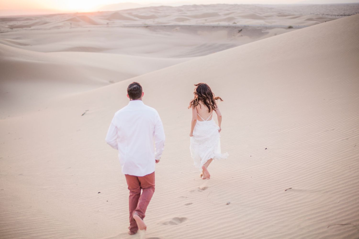 Couple runs playfully through sandy desert dunes in white attire.
