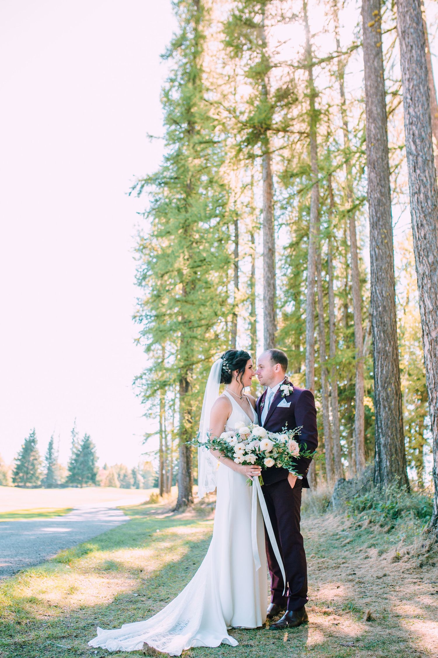A romantic wedding couple stands among tall pine trees in a sunlit forest setting holding a floral bouquet.