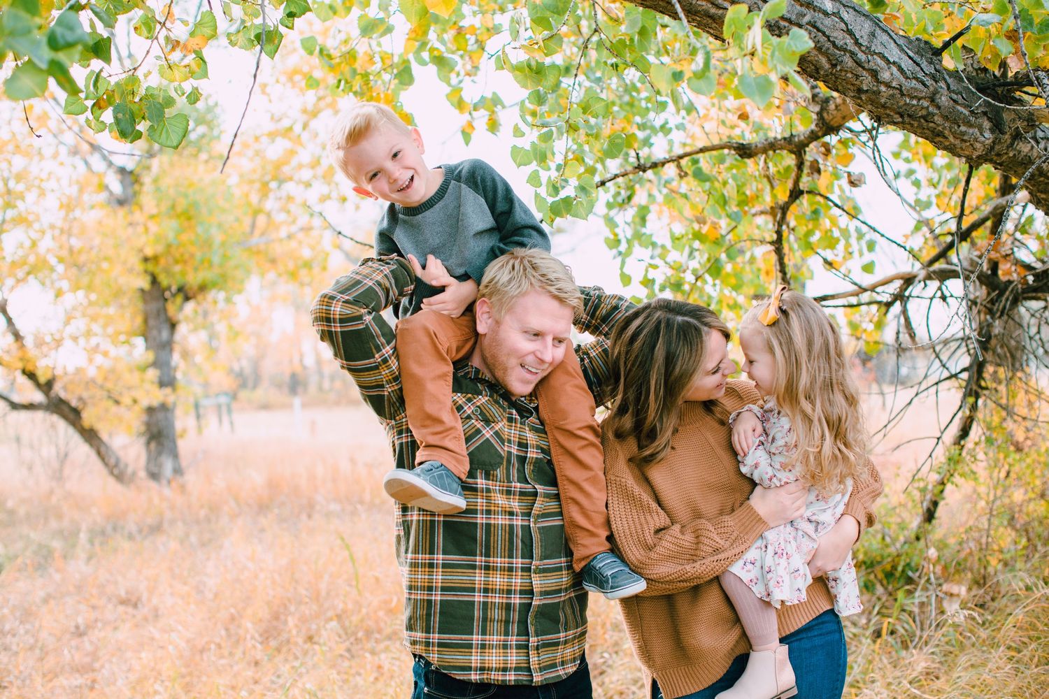 A family in coordinating autumn outfits poses together under yellow fall foliage in a park setting.