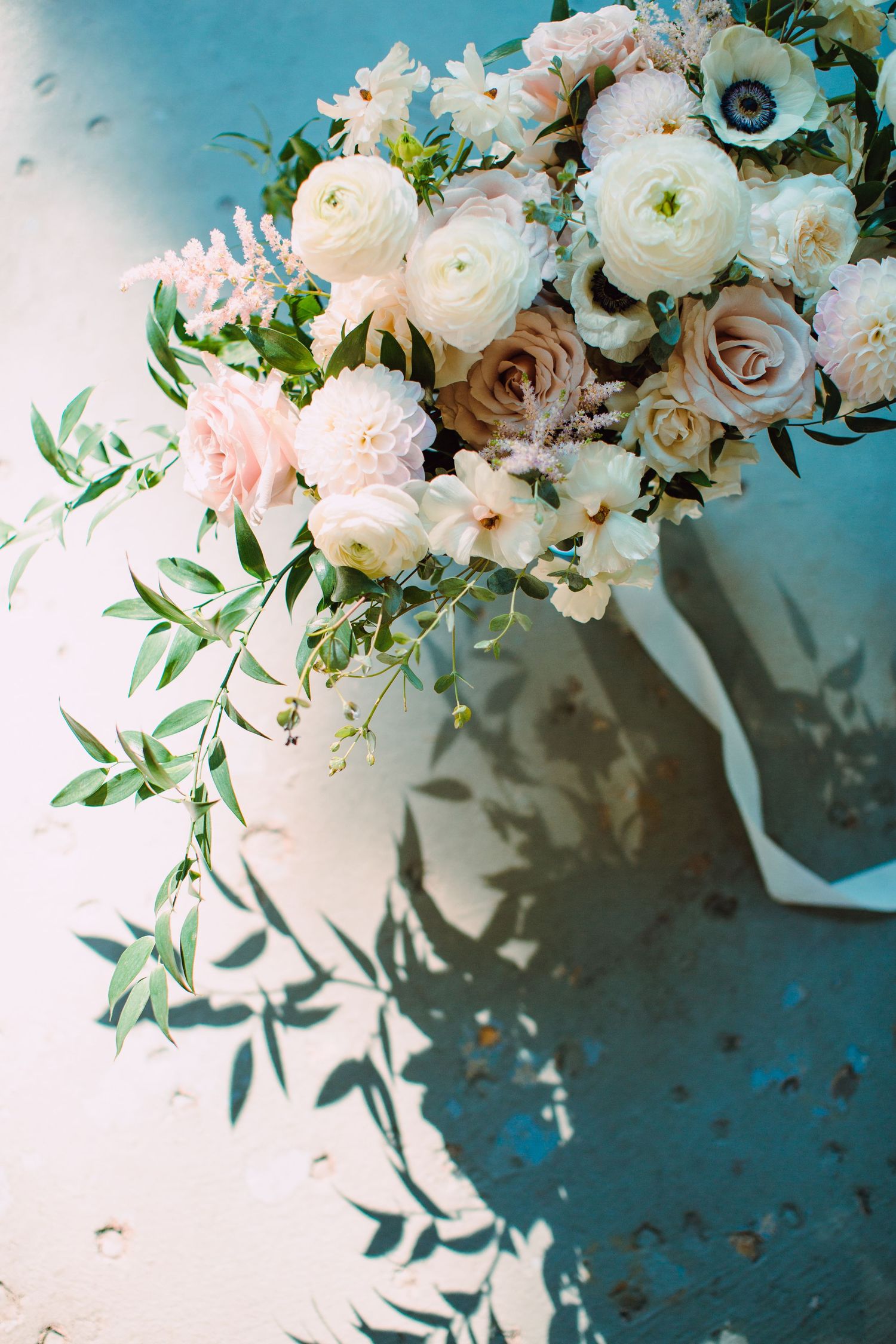 Elegant white ranunculus and blush roses arranged in a floral display against a moody teal blue background.