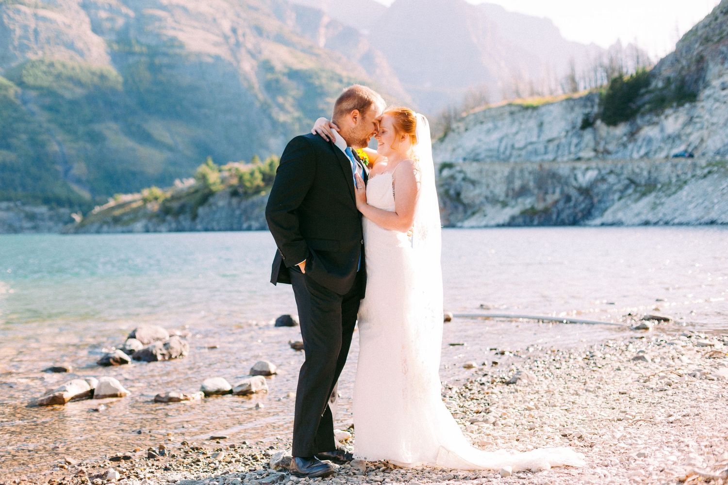 A romantic wedding couple shares an intimate moment on a rocky beach with majestic mountains and turquoise lake in background.