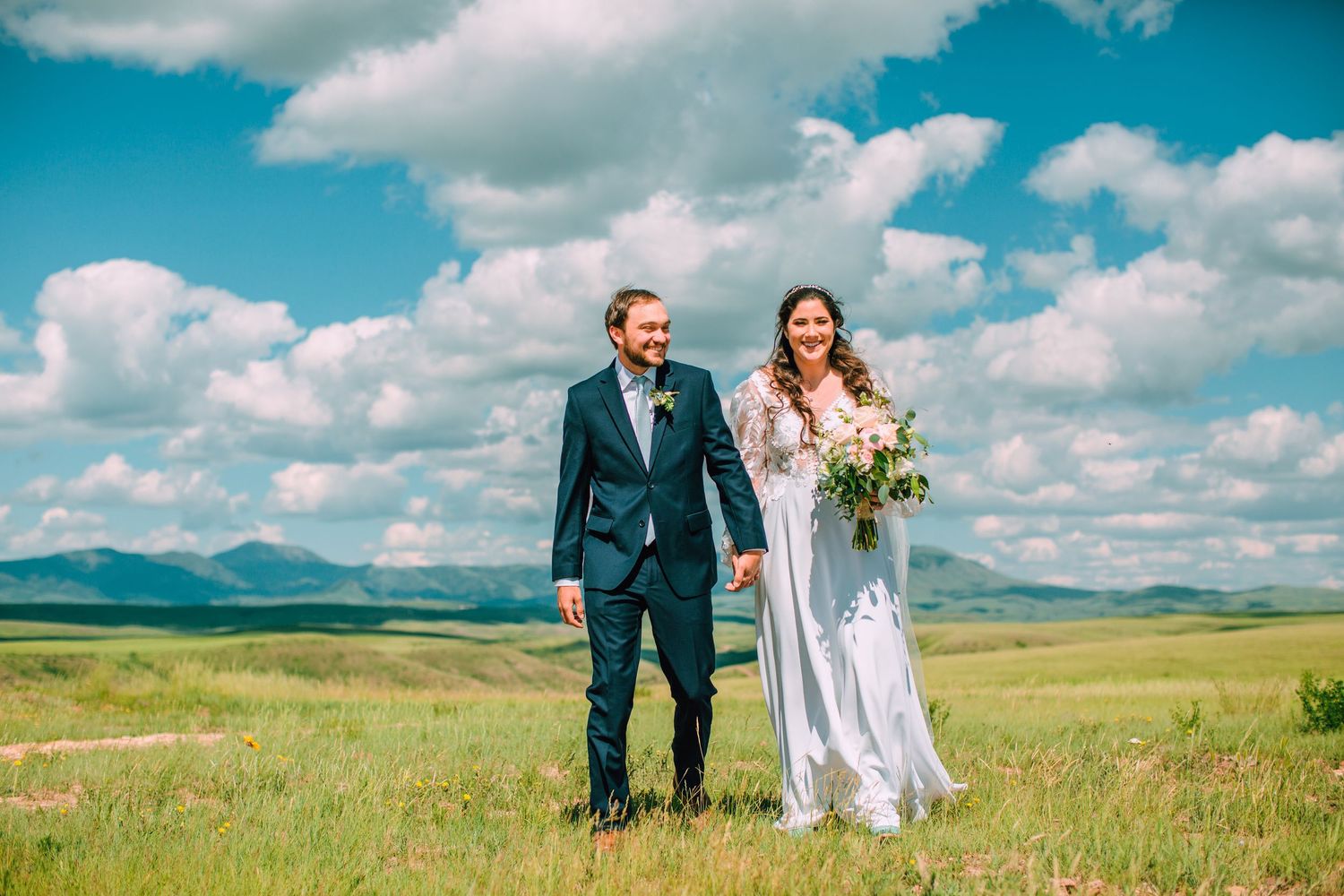 A couple in wedding attire walks through a scenic mountain meadow with dramatic clouds in the sky.