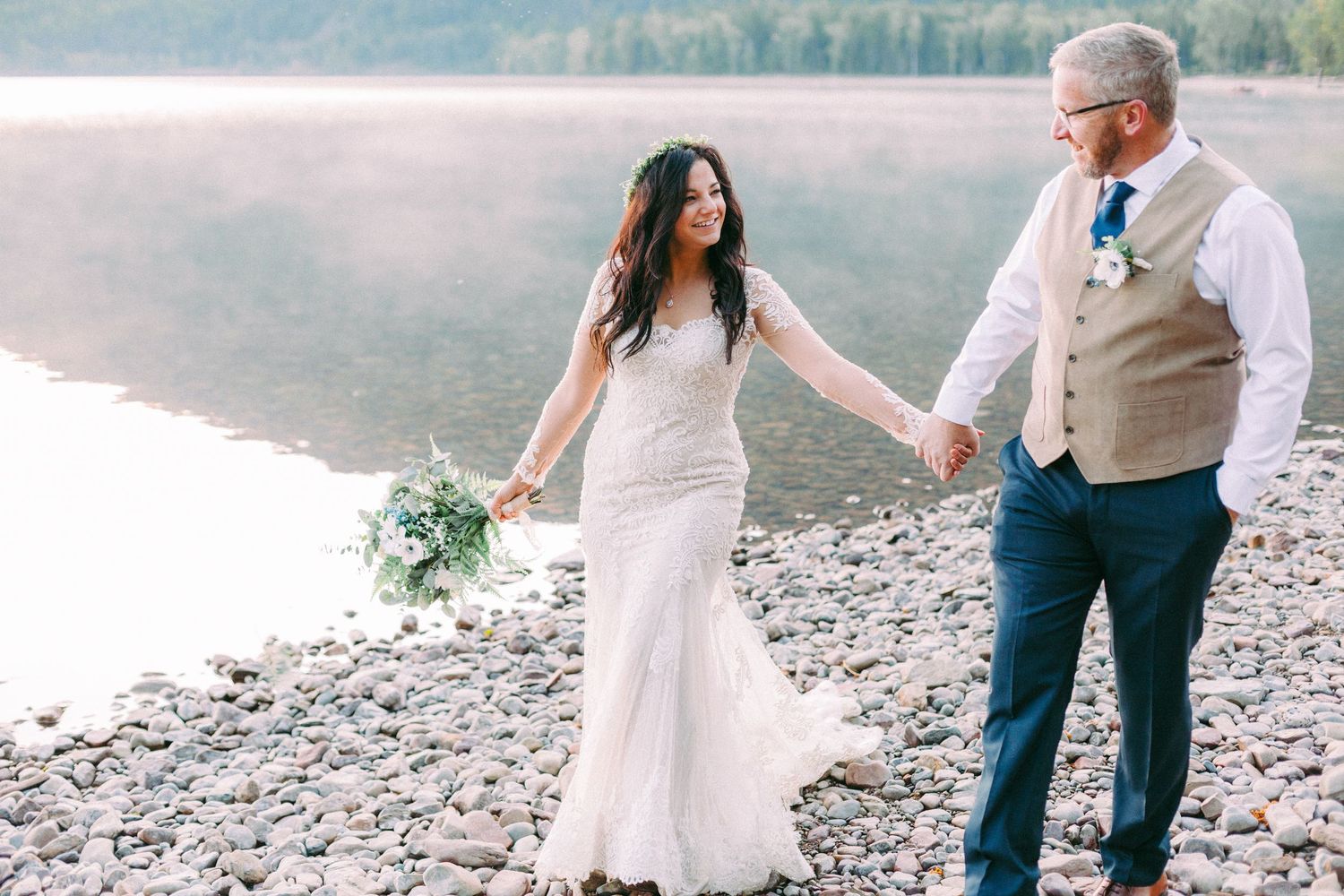 A couple in wedding attire walks hand in hand along a rocky lakeshore.