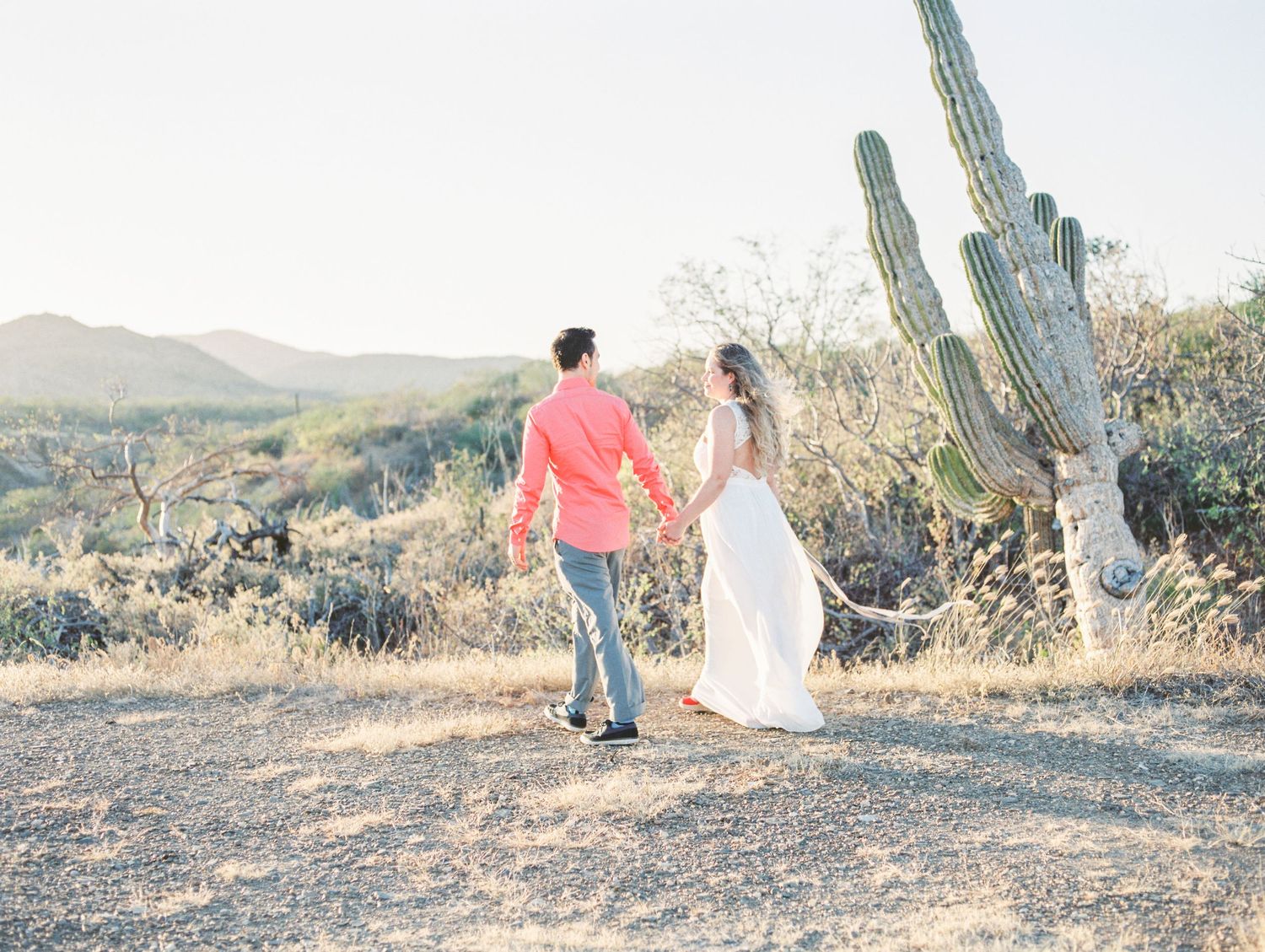 Couple walks together among desert cacti and mountain landscapes at sunset.