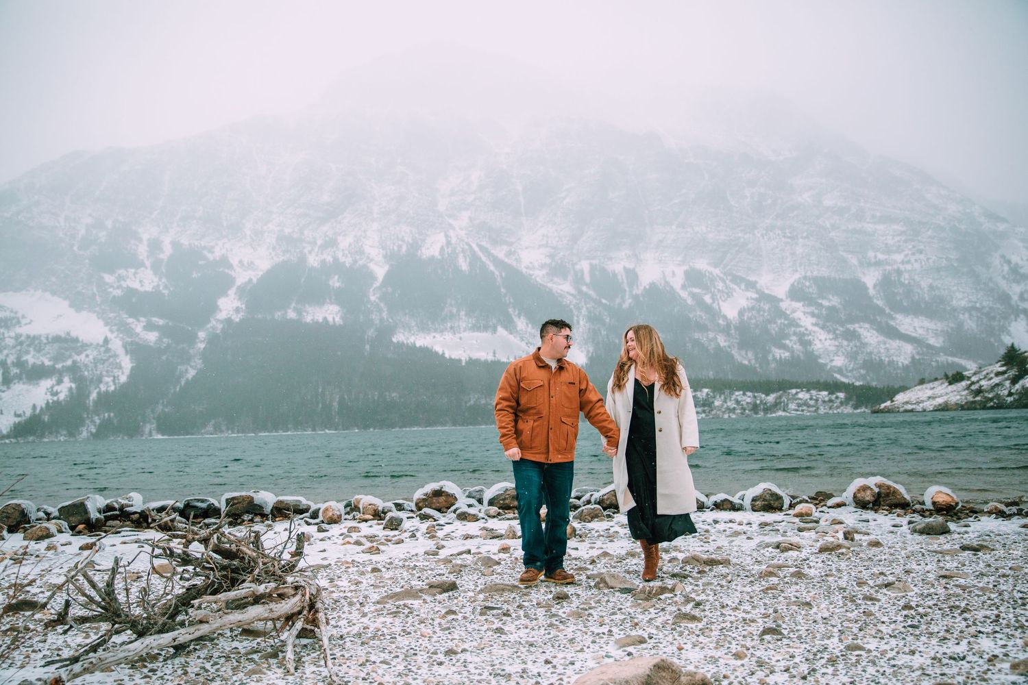 A couple stands on a rocky shore with snow-capped mountains creating a dramatic backdrop in winter.