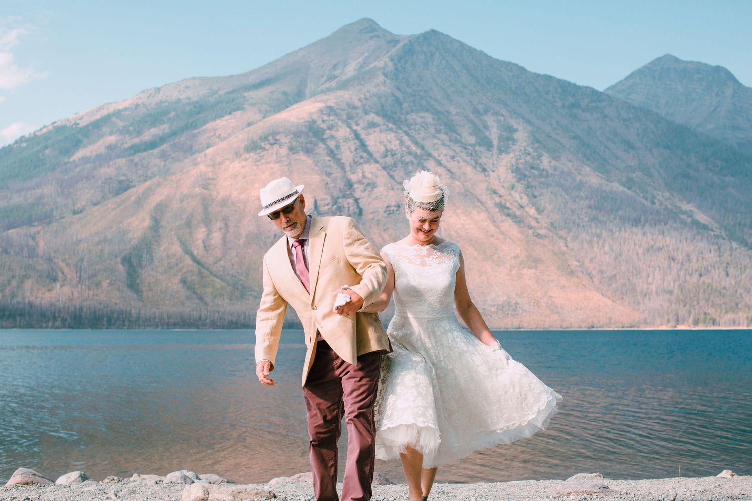 Romantic couple walks along mountain lake shoreline in wedding attire as veil flows in breeze during sunset photoshoot.