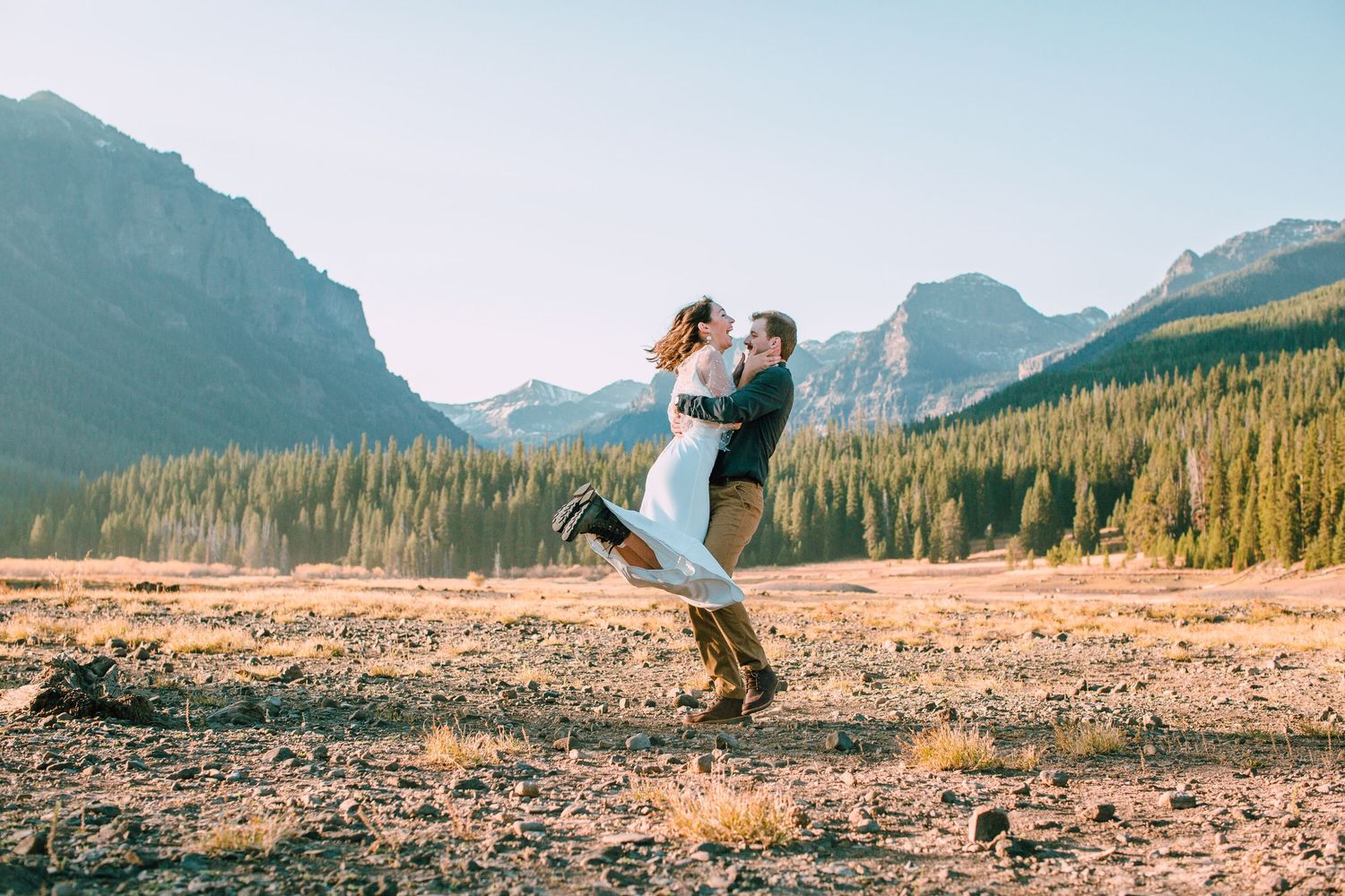 Couple embracing in a scenic mountain landscape with evergreen forests and dramatic peaks in the background.