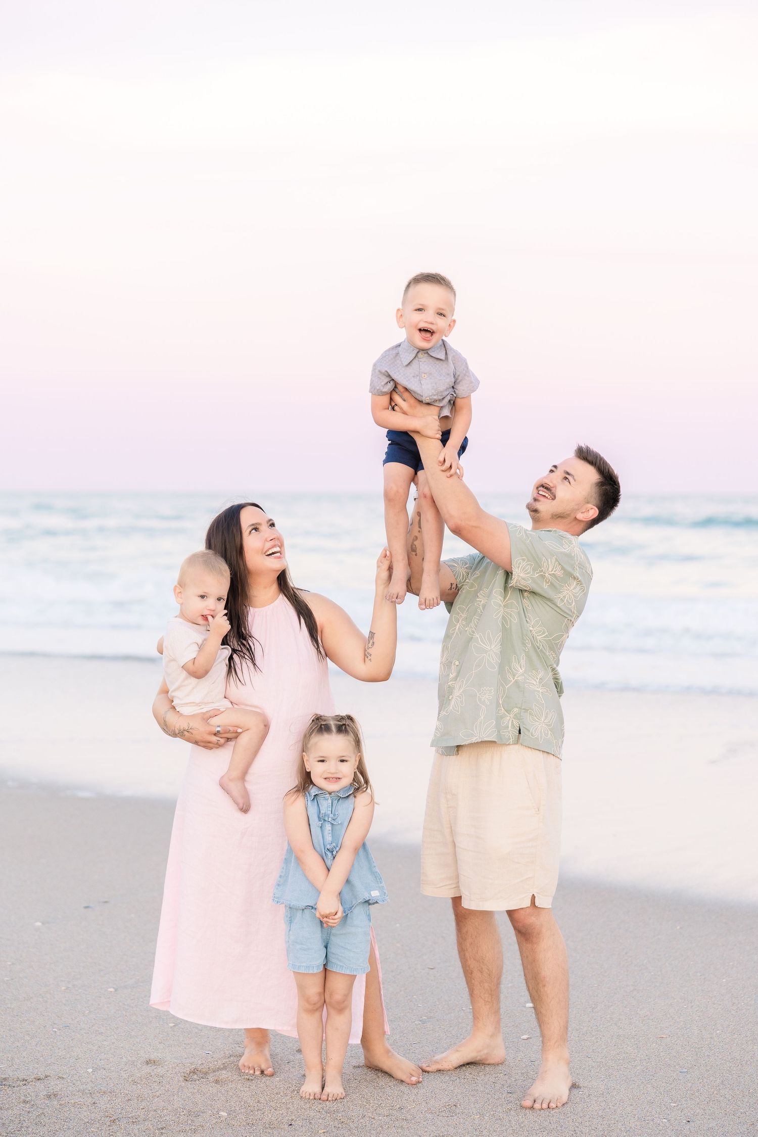 Happy family of five enjoying a beach sunset, parents lifting laughing children on sandy shore.