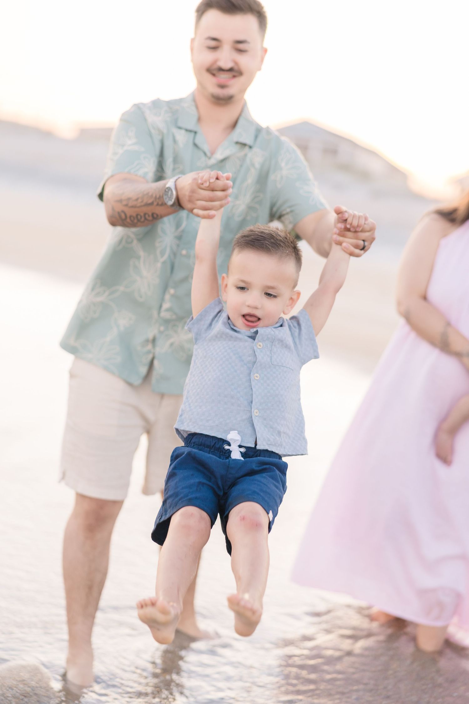 A father swings his young son over shallow beach water while a woman in a pink dress stands nearby at sunset.
