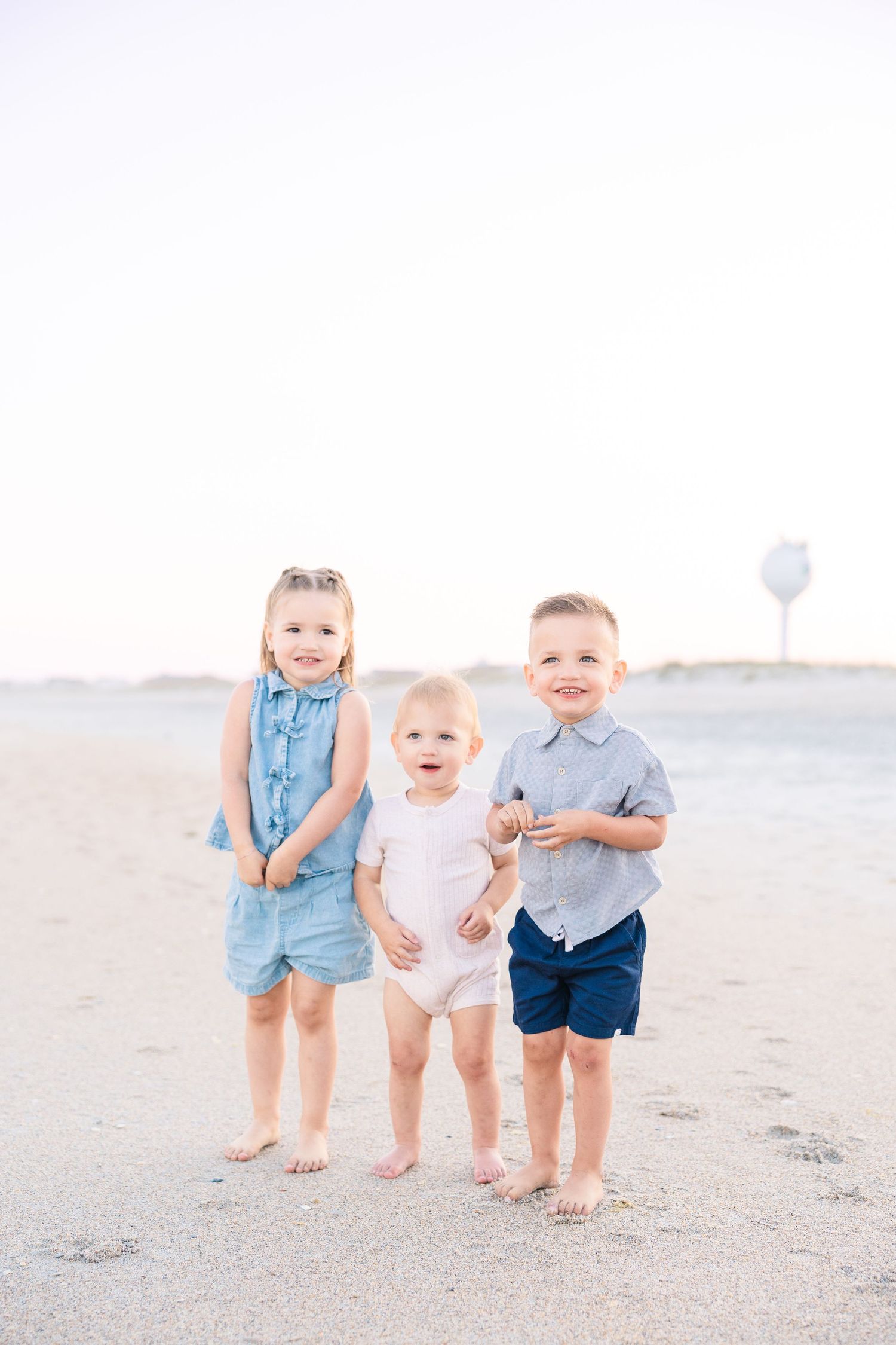 Three young siblings stand barefoot on a sandy beach, two toddlers flanking a smiling baby in white romper.