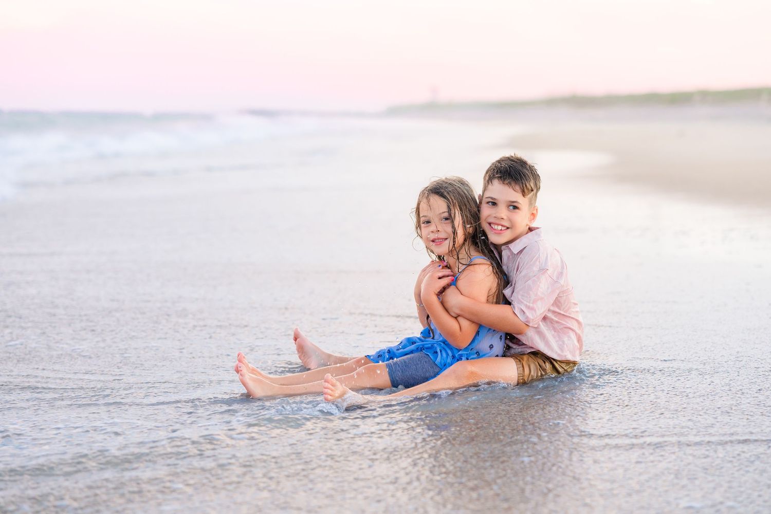 Two children sitting together in shallow ocean waves on sandy beach.