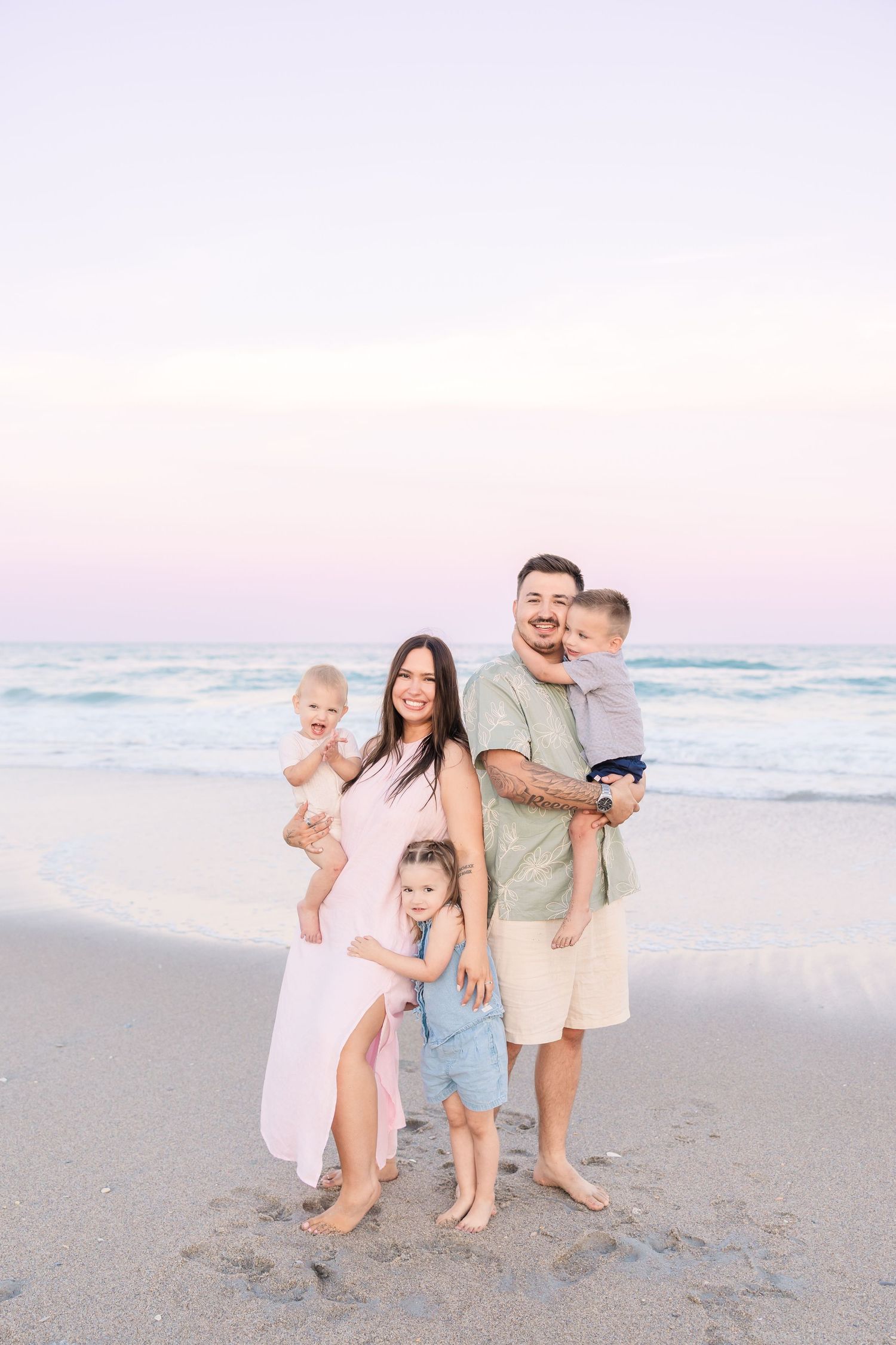 A smiling family of five stands together on a beach at dusk with pastel pink sky behind.