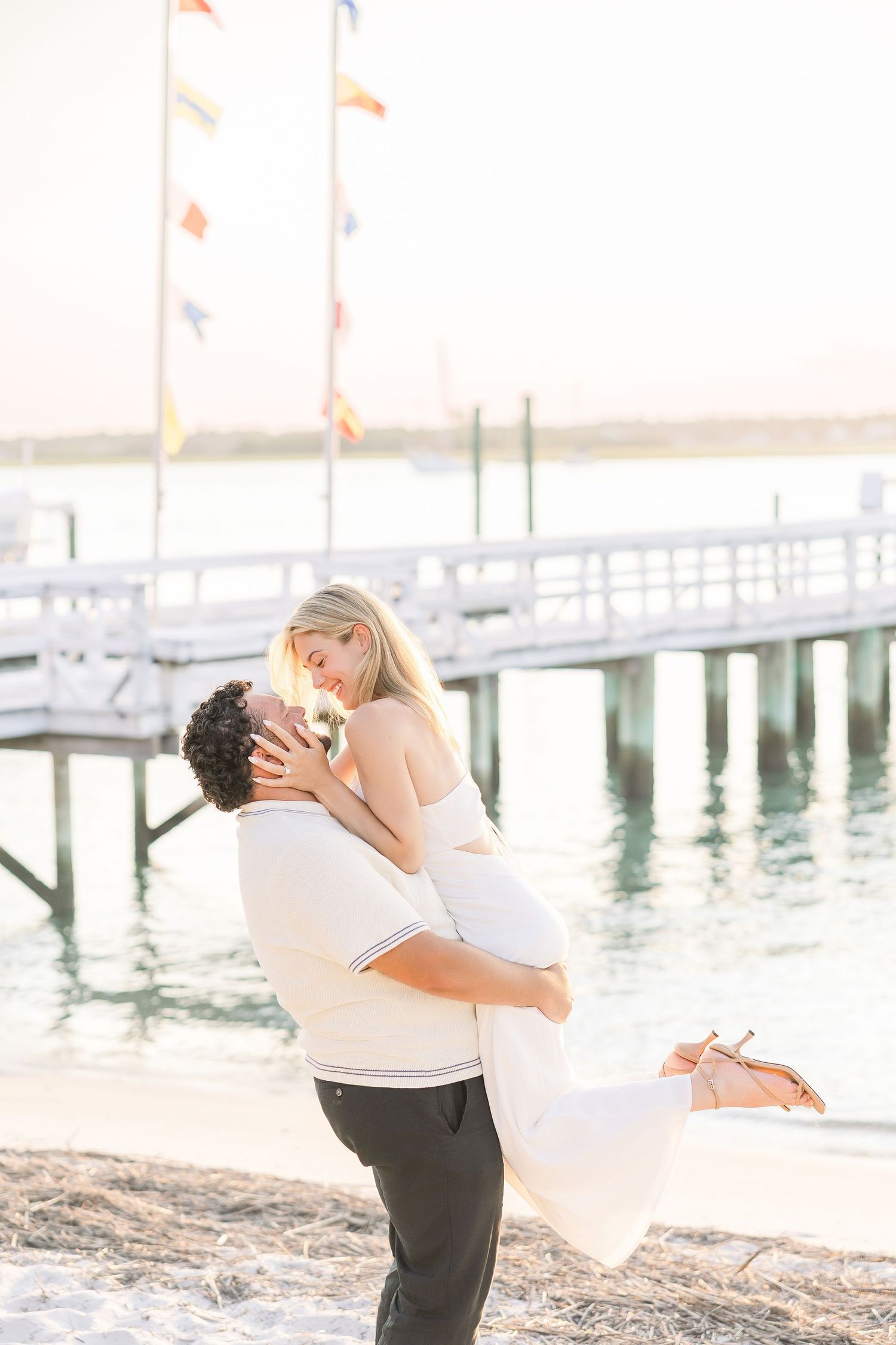 Man lifting woman in white dress on sandy beach near pier, colorful flags in background at sunset.