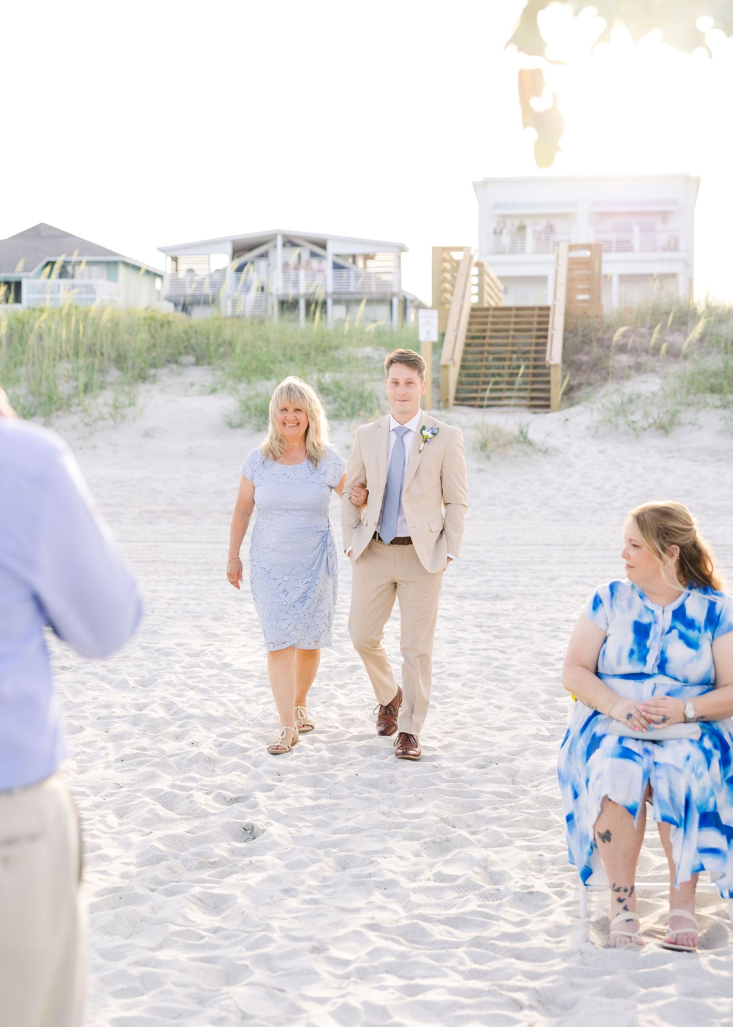 Groom in tan suit walks with woman in blue dress down sandy beach aisle at outdoor wedding ceremony in Carolina Beach near Wilmington, NC.