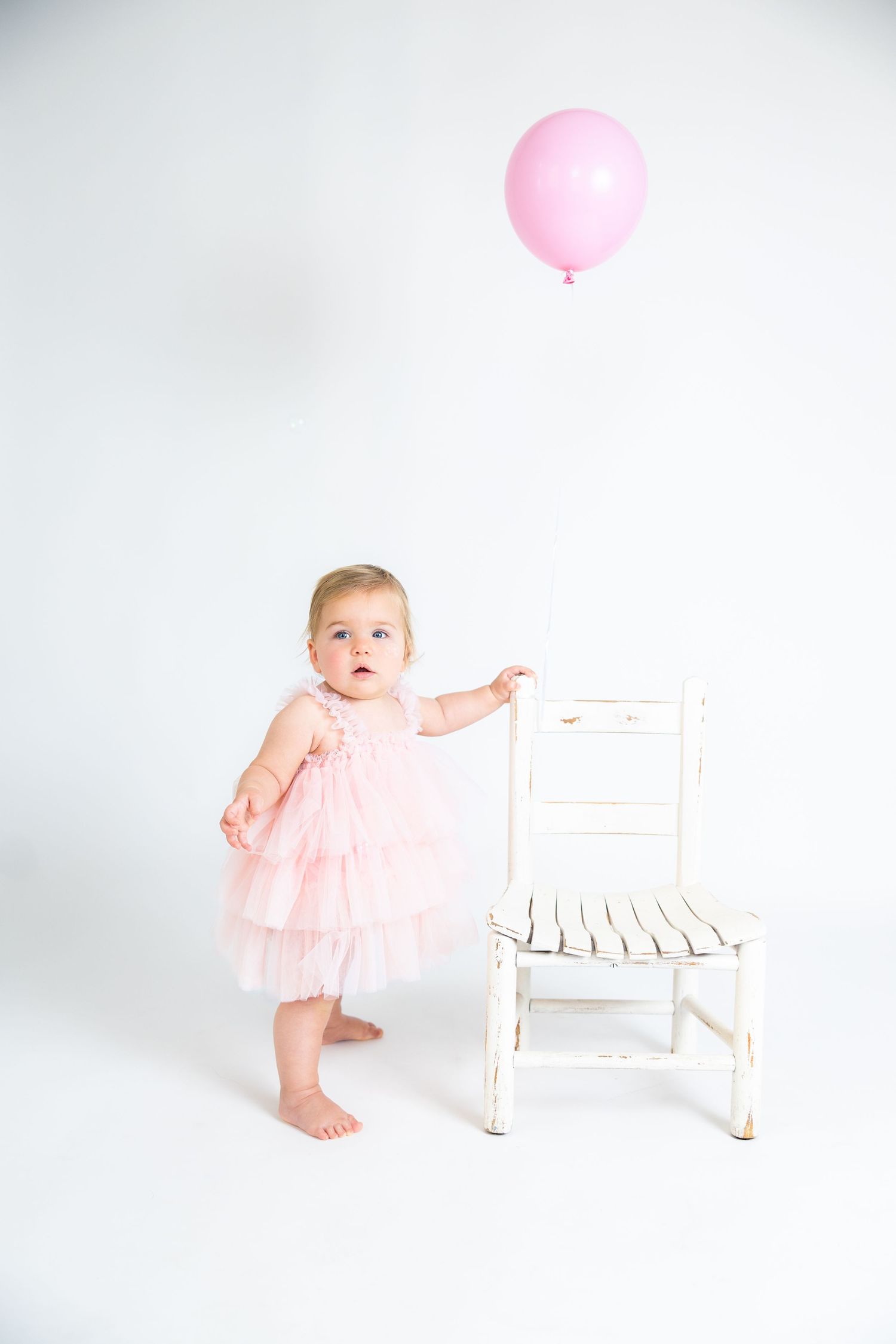 Baby in pink tulle dress standing next to white chair with pink balloon.