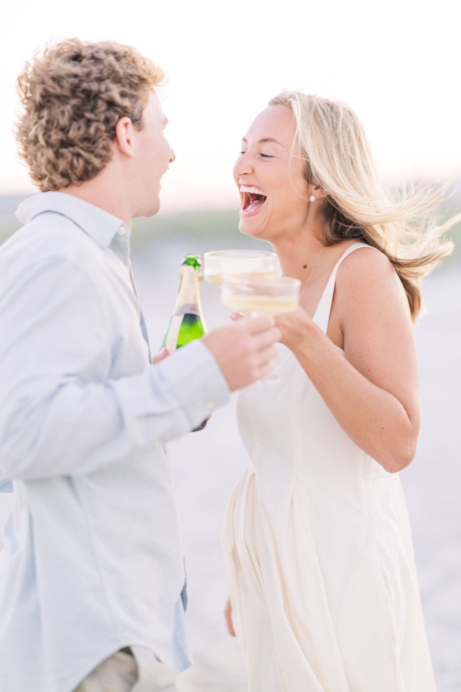 Joyful couple laughs while sharing champagne on a sunlit beach in casual elegant attire.