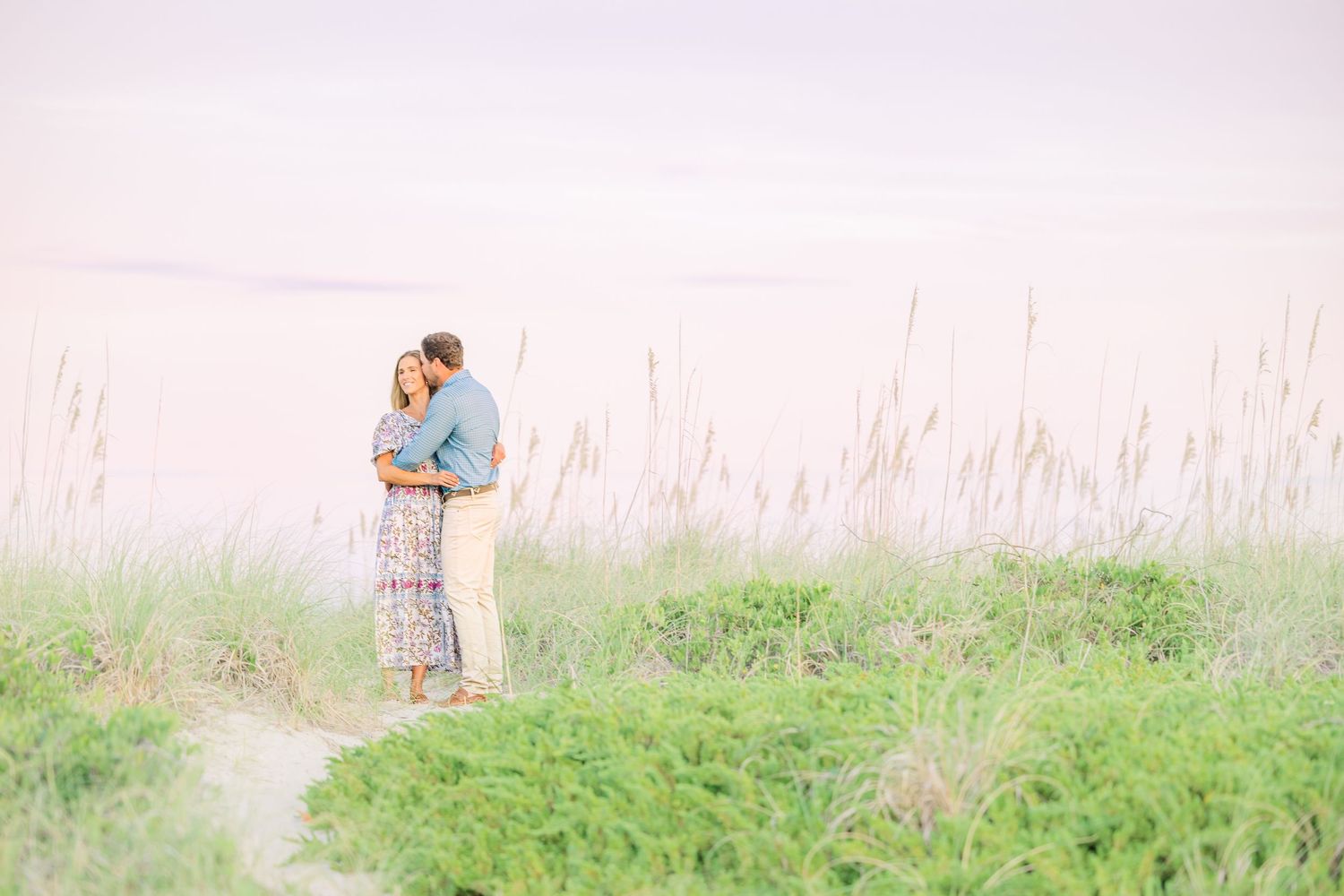A couple shares an intimate moment among sea oats during a pastel sunset.