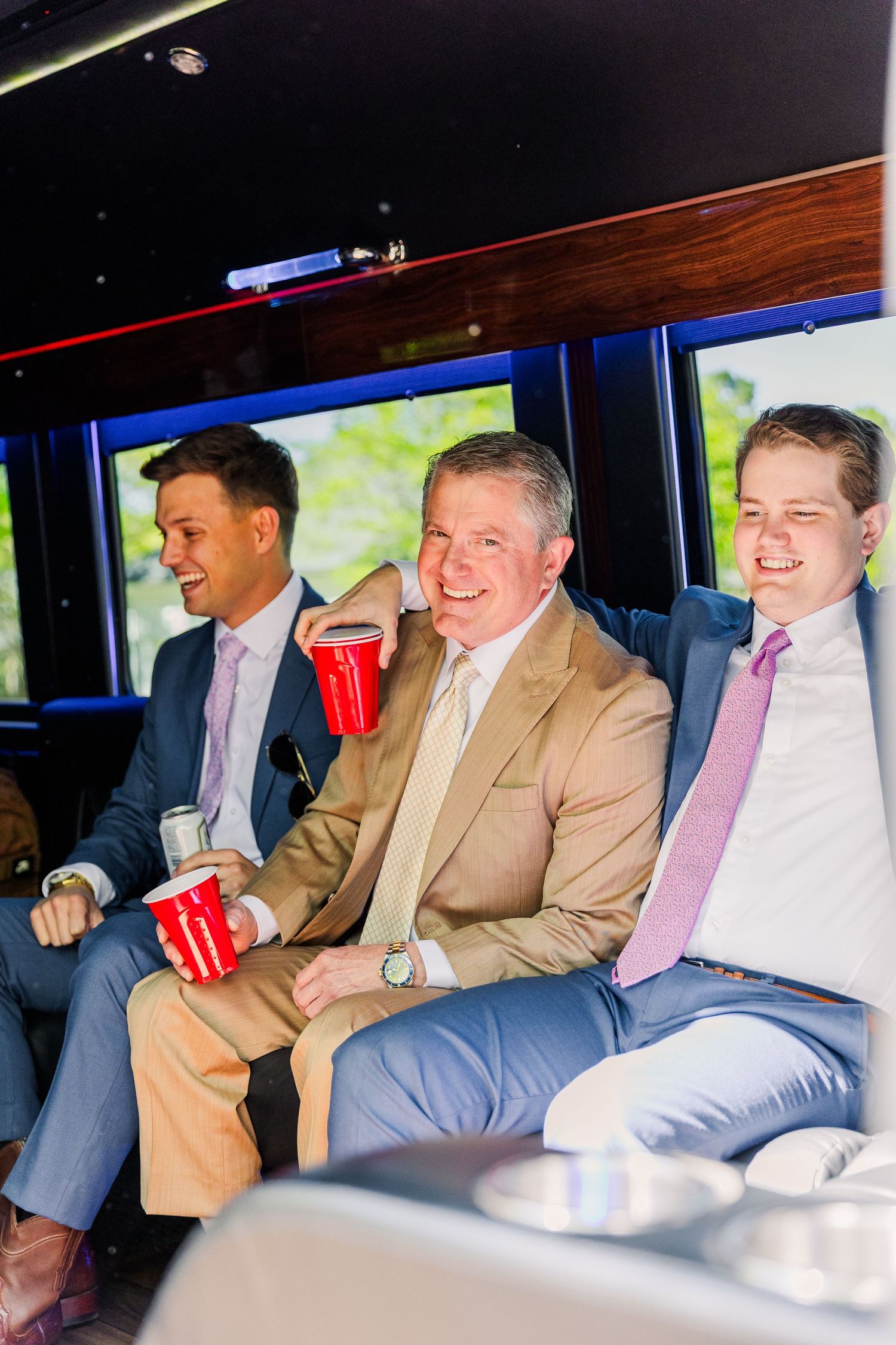 Three men in suits share drinks and laughs while riding in a luxury vehicle.