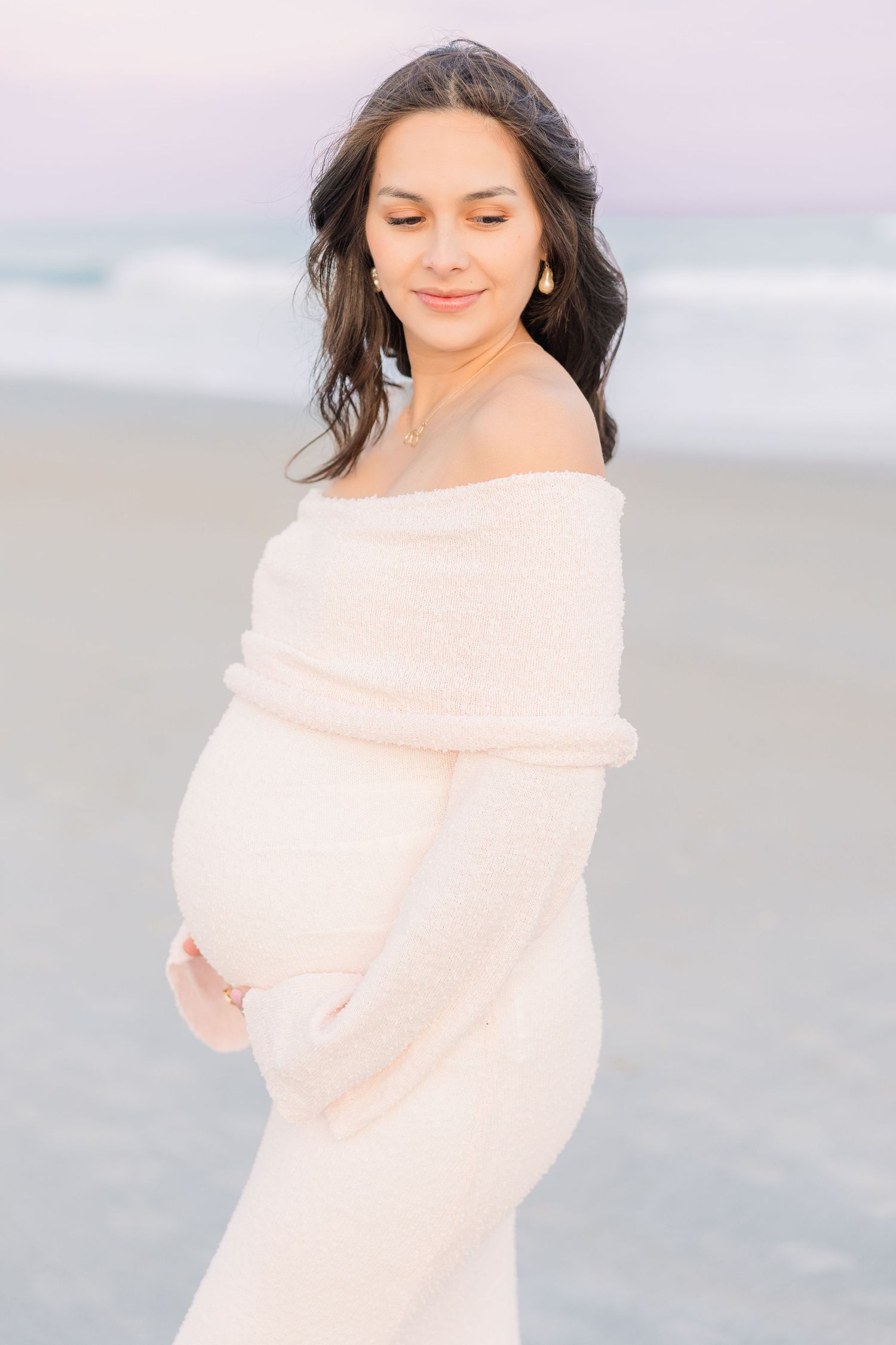 Woman in off-shoulder blush sweater poses elegantly on beach at sunset.