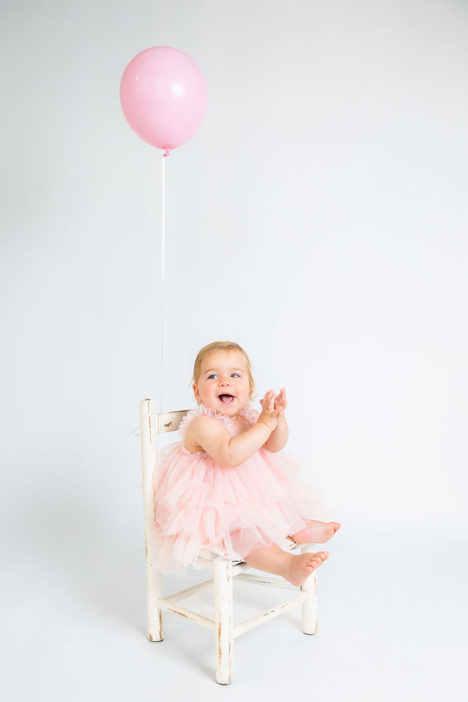 Baby in pink dress sitting on white chair with pink balloon against white backdrop.