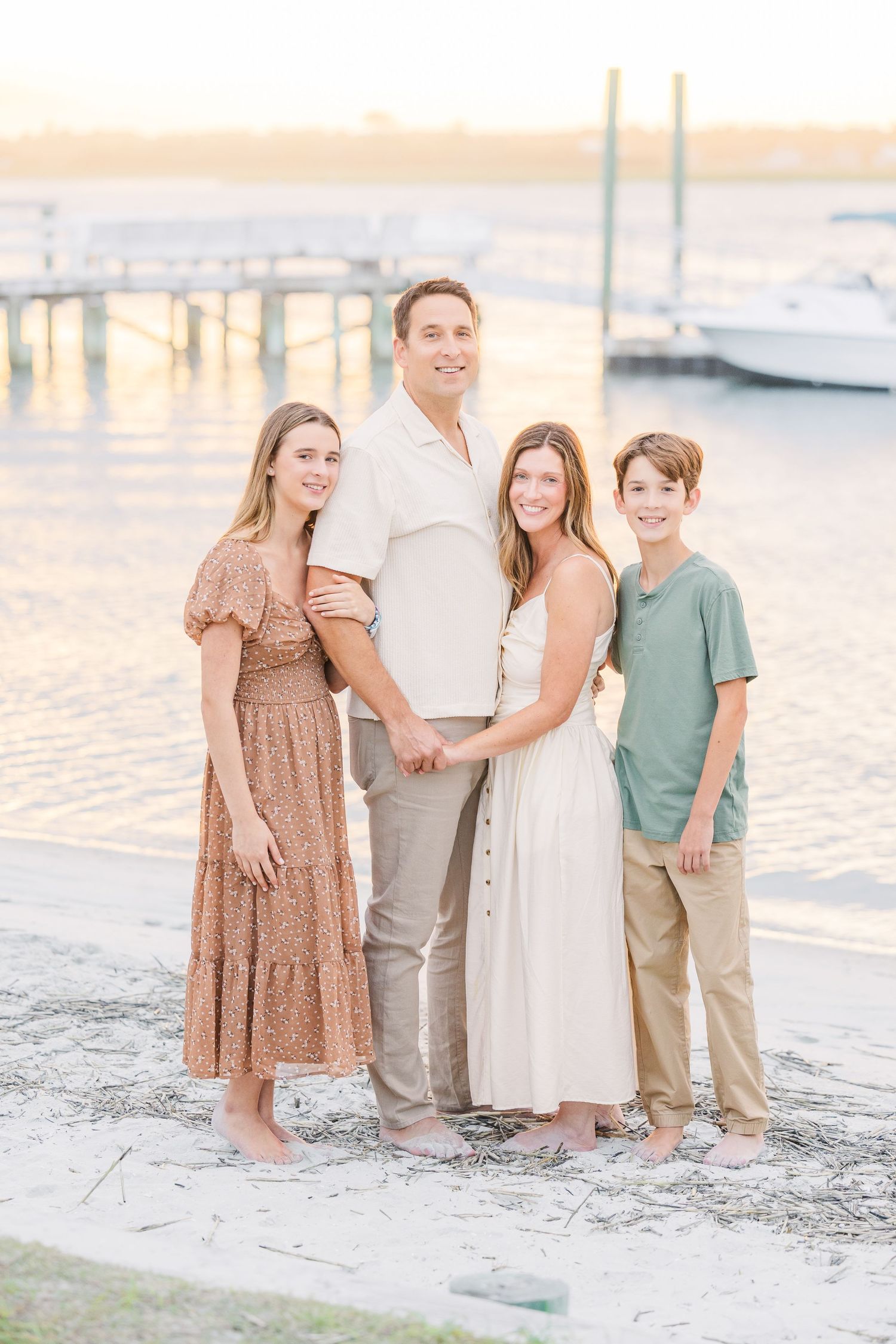 Family wearing coordinated neutral outfits poses together on sandy beach at sunset.