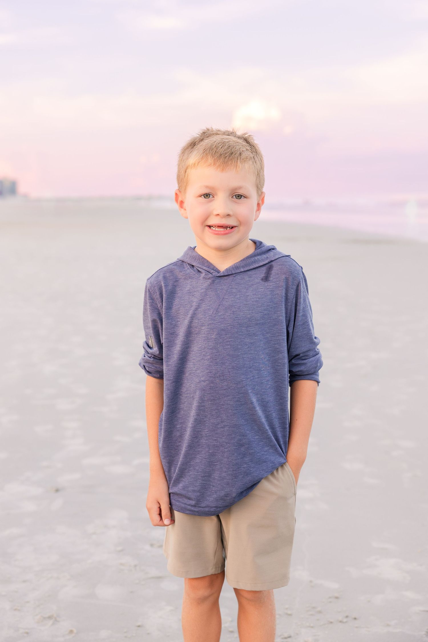 Young child in blue hoodie and khaki shorts playing on beach during sunset.
