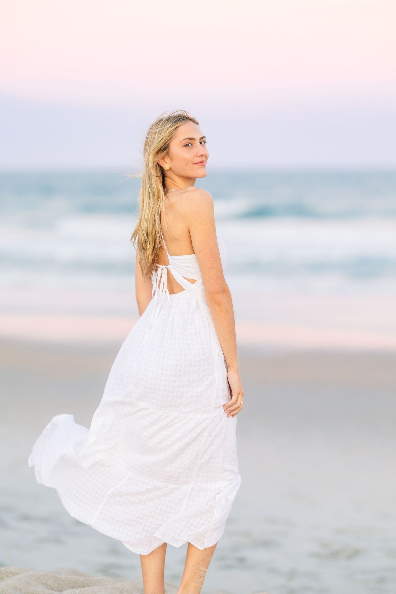 A person poses in a white tie-back dress on the beach during sunset with waves rolling in.