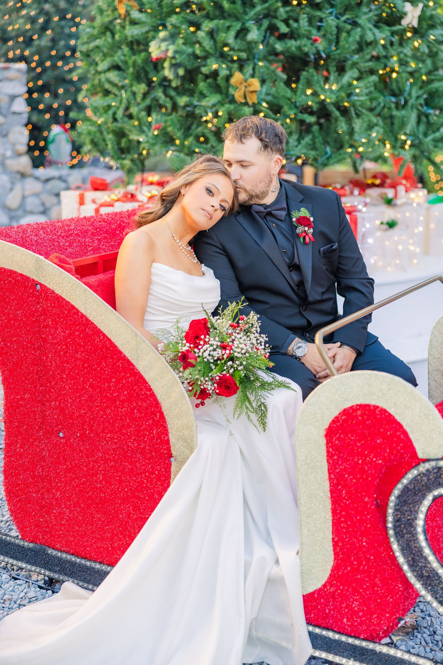 Romantic couple poses in red holiday sleigh with Christmas trees and twinkling lights.