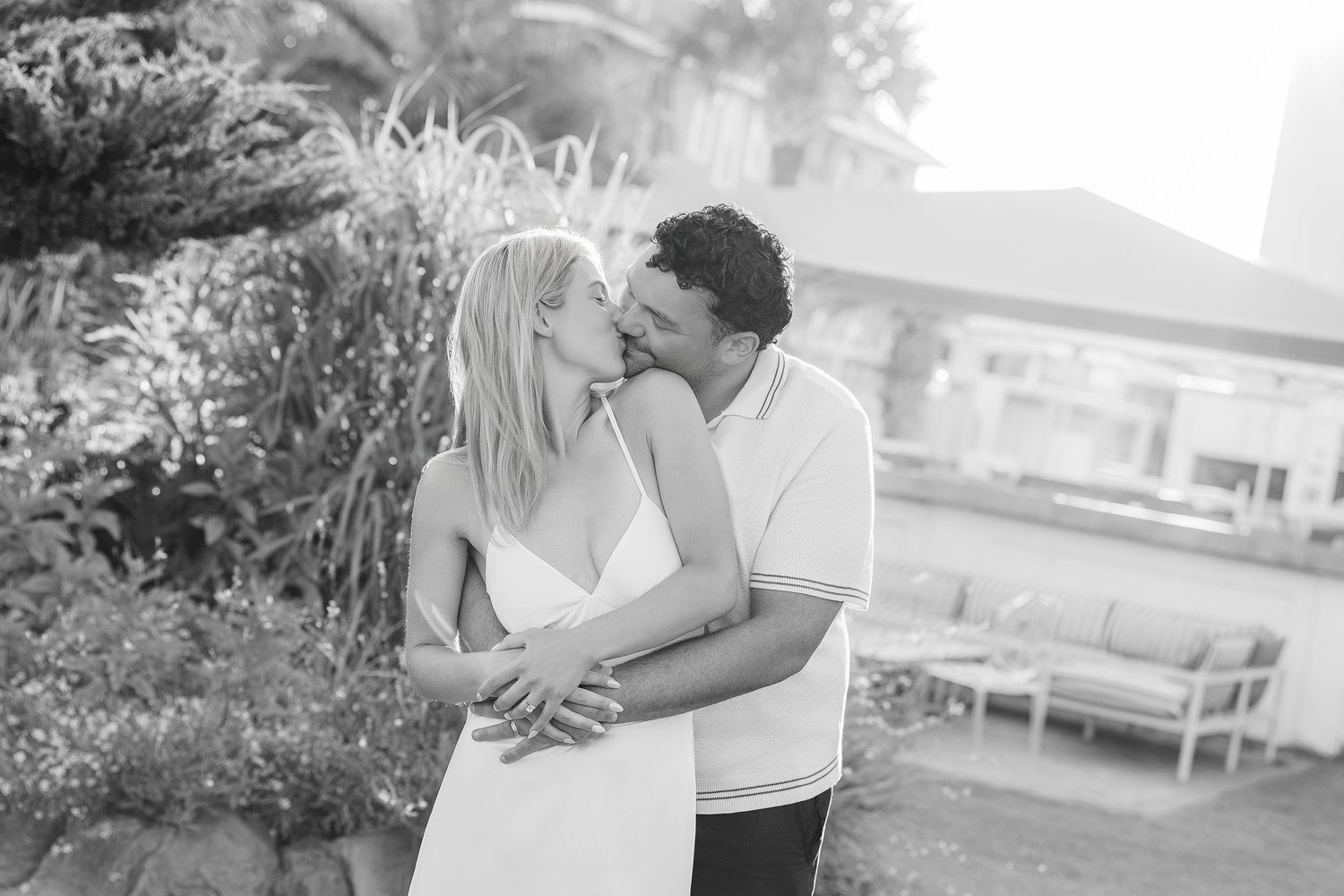 Black and white photo of a couple sharing a kiss outdoors, surrounded by lush greenery.