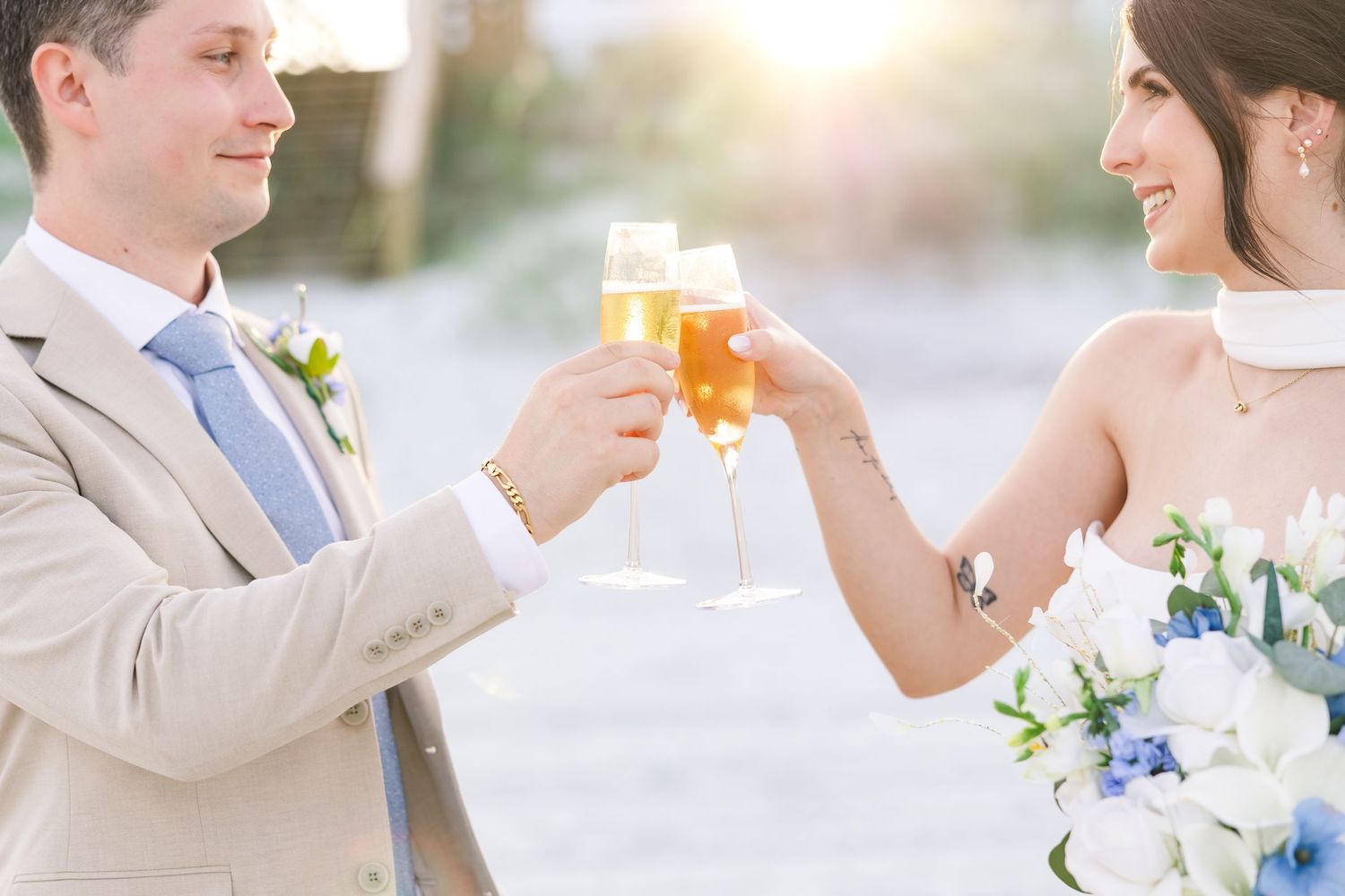 Bride and groom toast with champagne glasses, she holds a blue and white floral bouquet on the beach in Carolina Beach near Wilmington, NC.