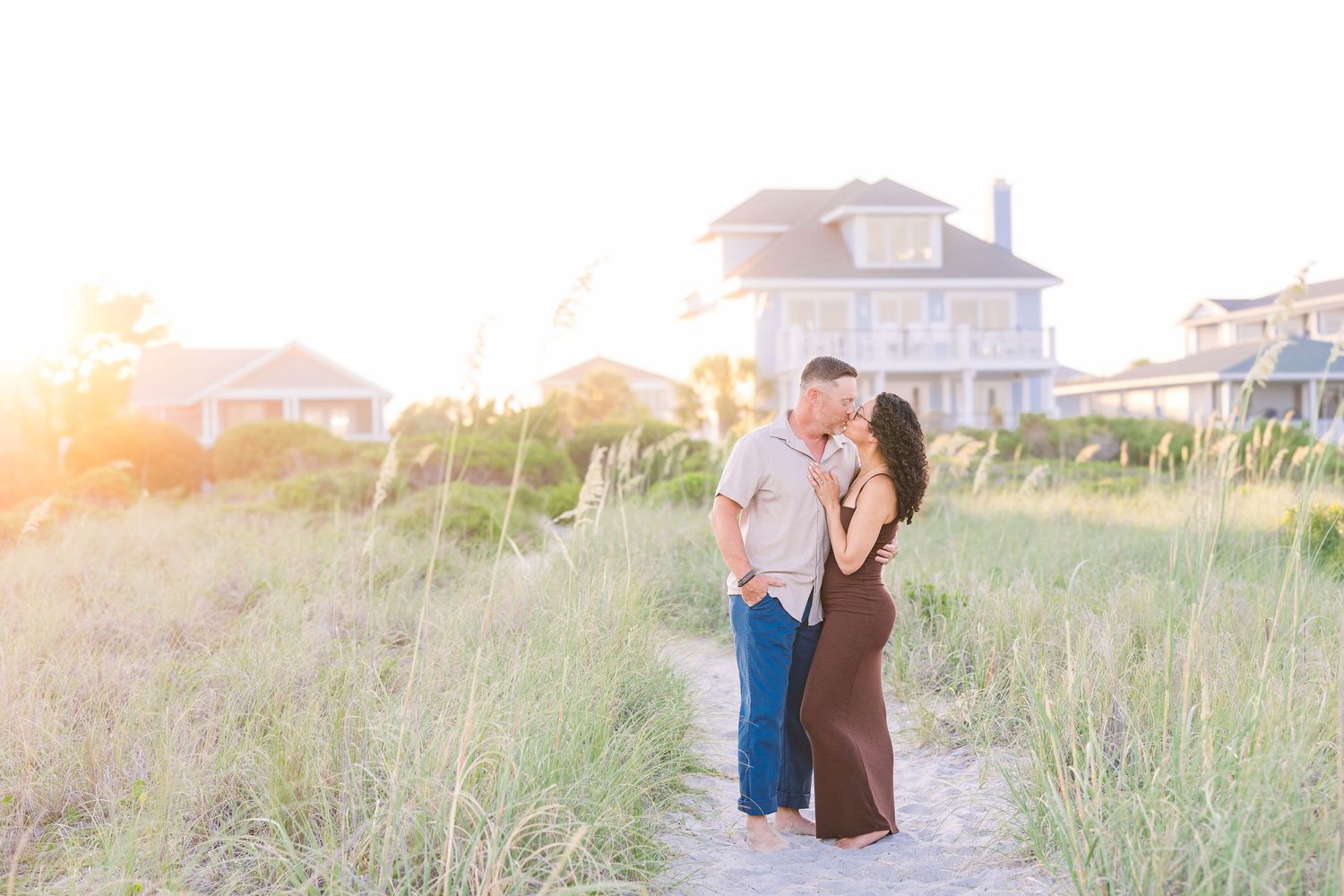 Couple sharing a romantic kiss on a sandy beach path surrounded by tall grass at golden hour sunset.