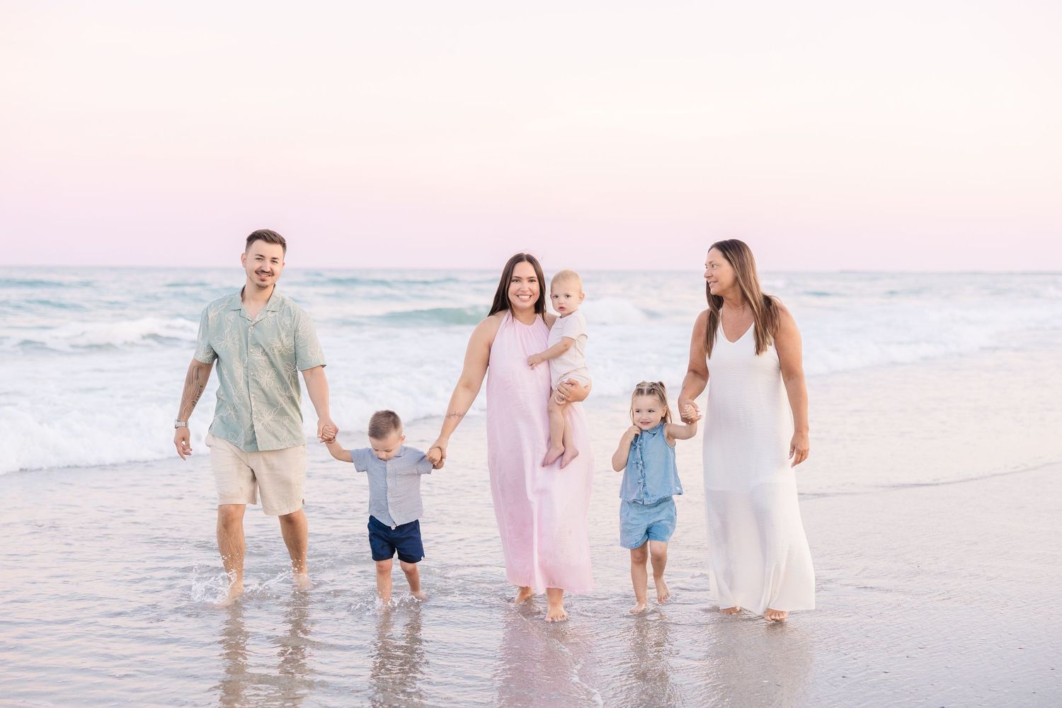 Family of five walking hand-in-hand along a beach at sunset with pastel pink sky and ocean waves.