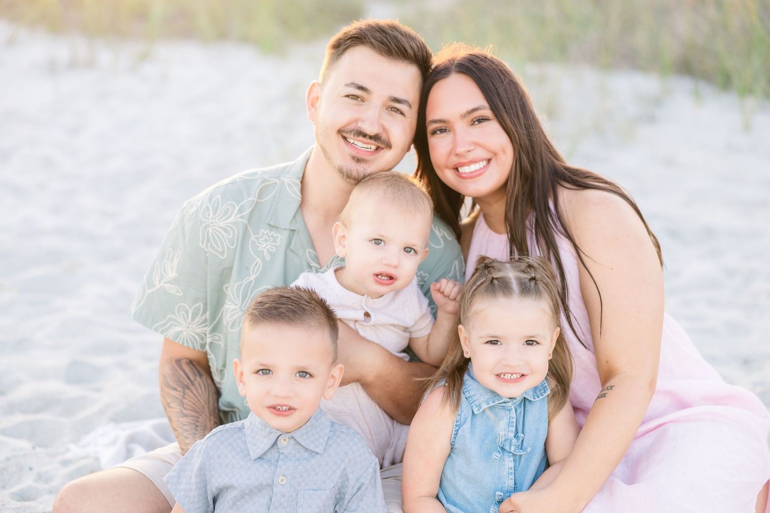 Happy family of five smiling together on a sandy beach, with parents holding three young children on a bright sunny day.