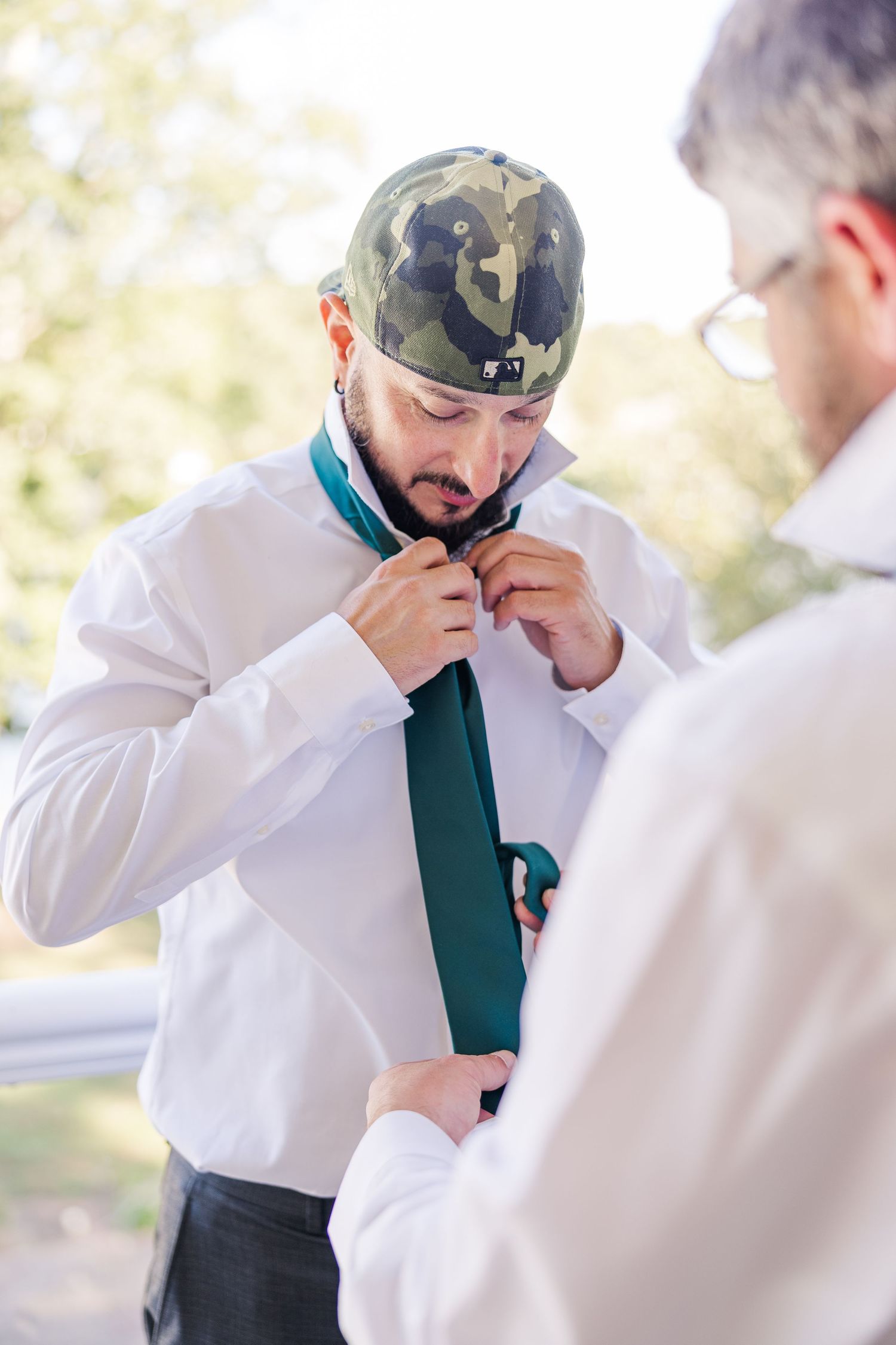 A person wearing a camo hat adjusts their tie with help from others wedding groom bride lake Gaston photographer light airy