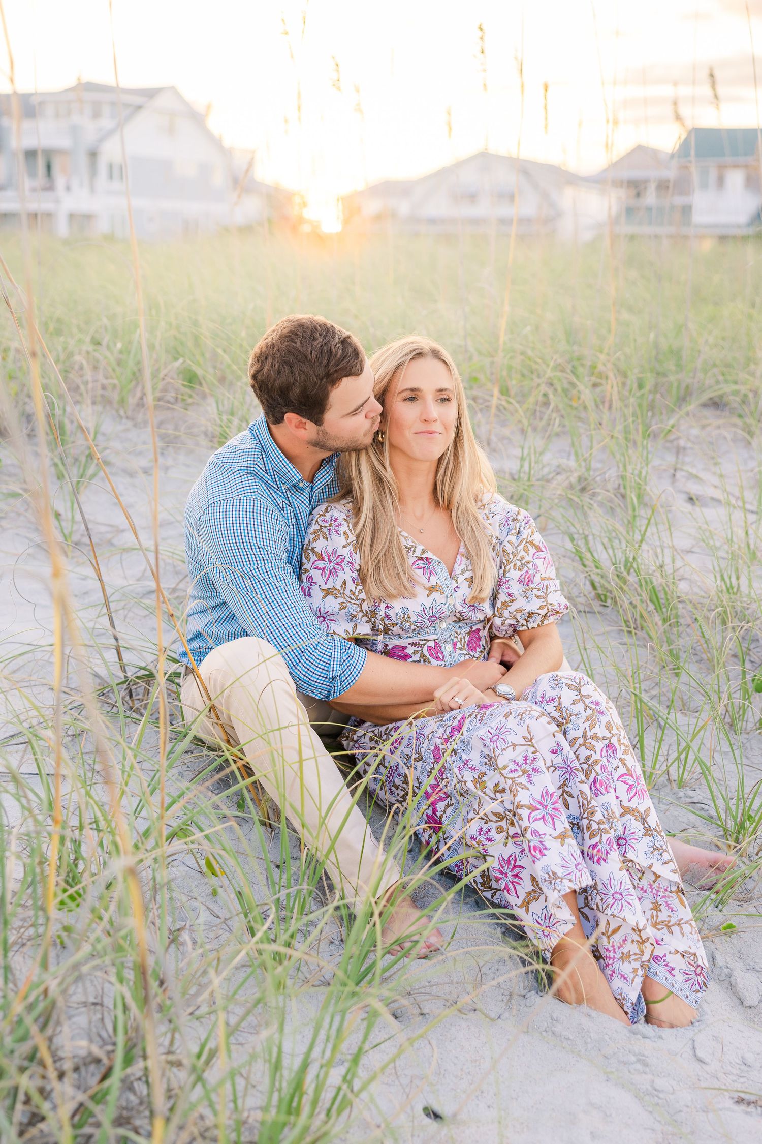 Couple sitting in beach grass at sunset with beach houses in background.