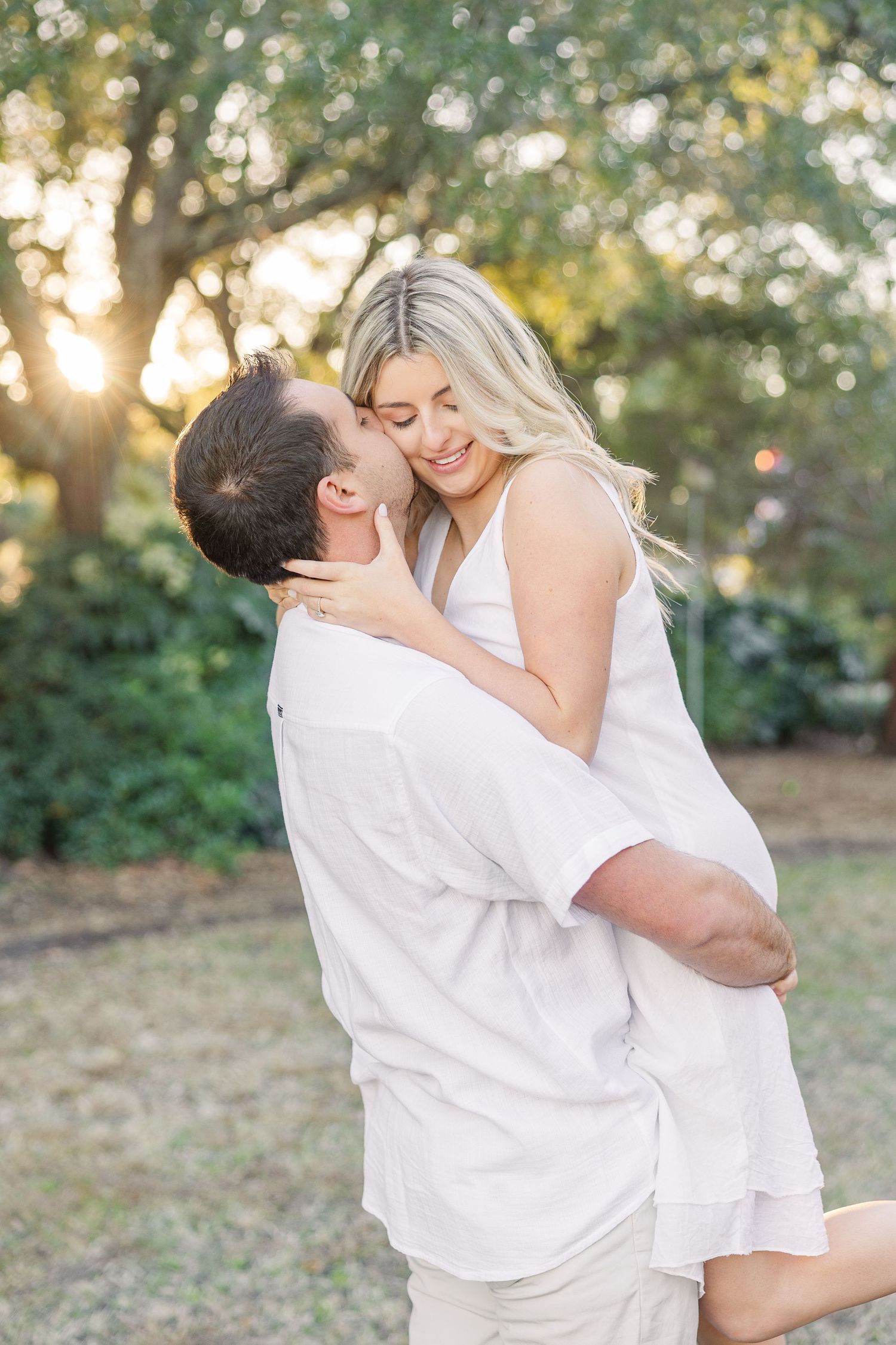 Two people dressed in white embrace and dance playfully in a park during golden hour.