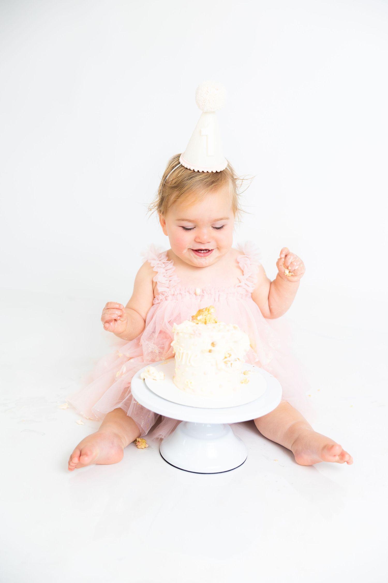 Baby wearing pink dress and party hat looking down at birthday cake during cake smash photo session.