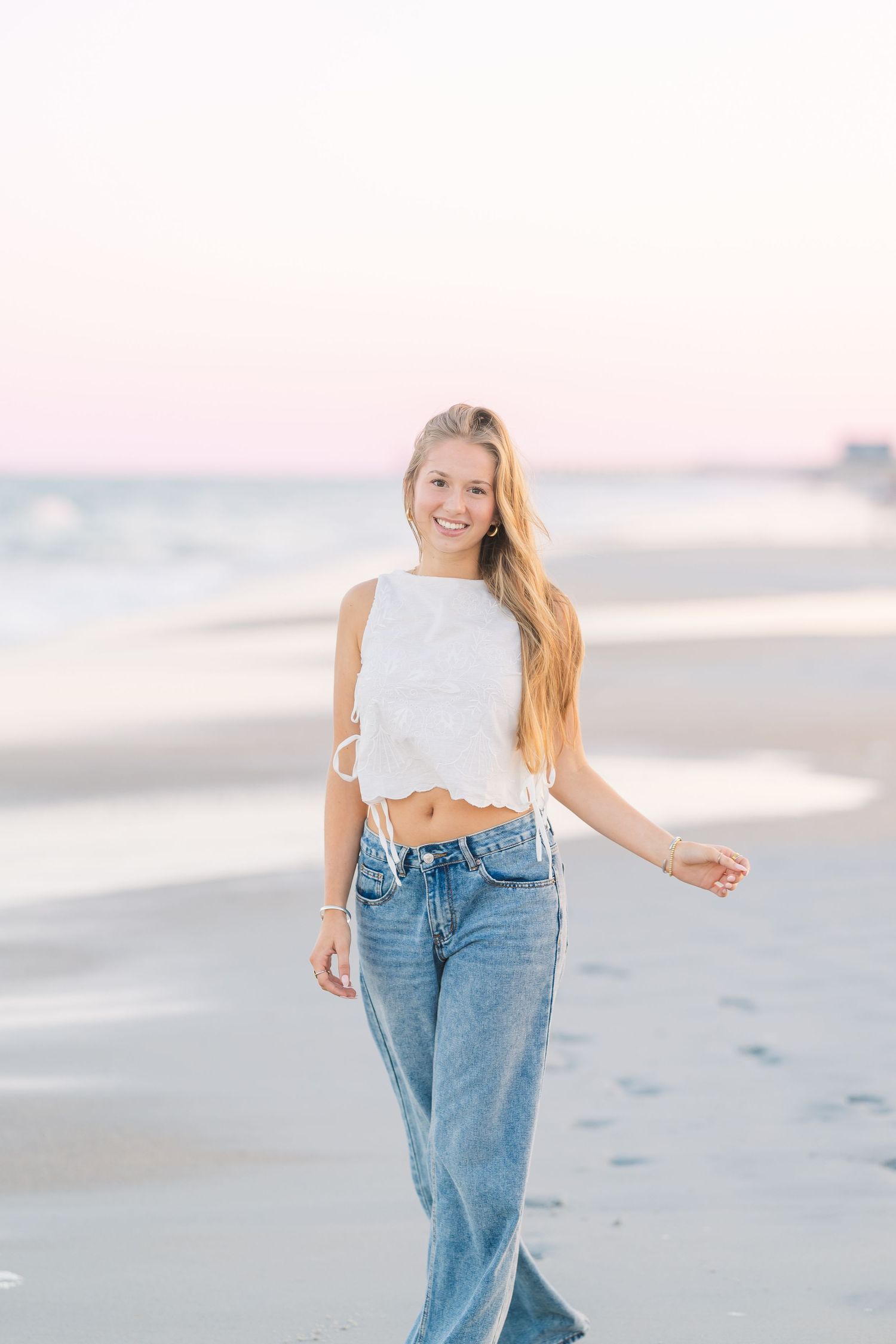 Woman in casual outfit walking and posing playfully on beach during sunset photoshoot.