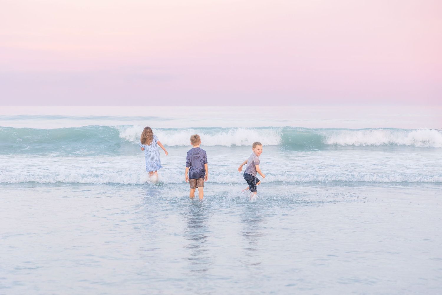 Three people playing and splashing in the ocean waves during a dreamy pink sunset.