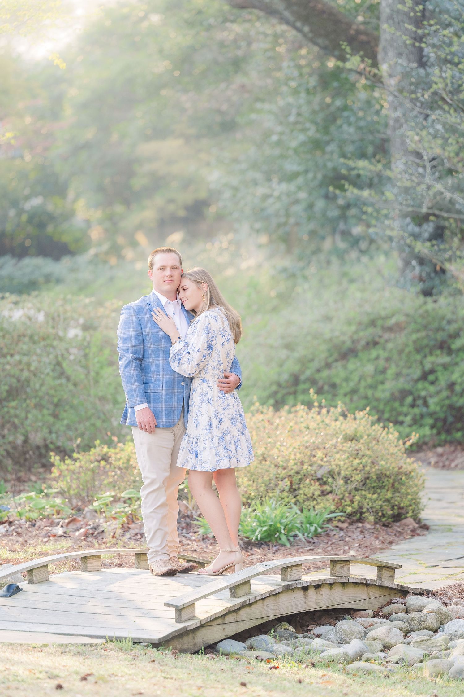 A couple stands together on a rustic wooden footbridge surrounded by lush greenery in an outdoor setting.