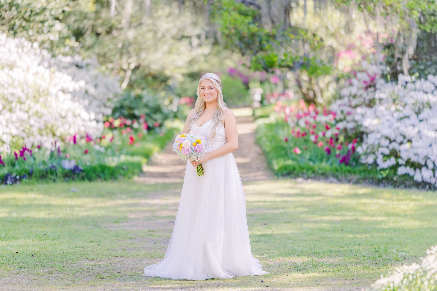 Bride in white gown holding pink bouquet stands in blooming garden path with colorful tulips and flowering trees in Airlie Gardens in Wilmin